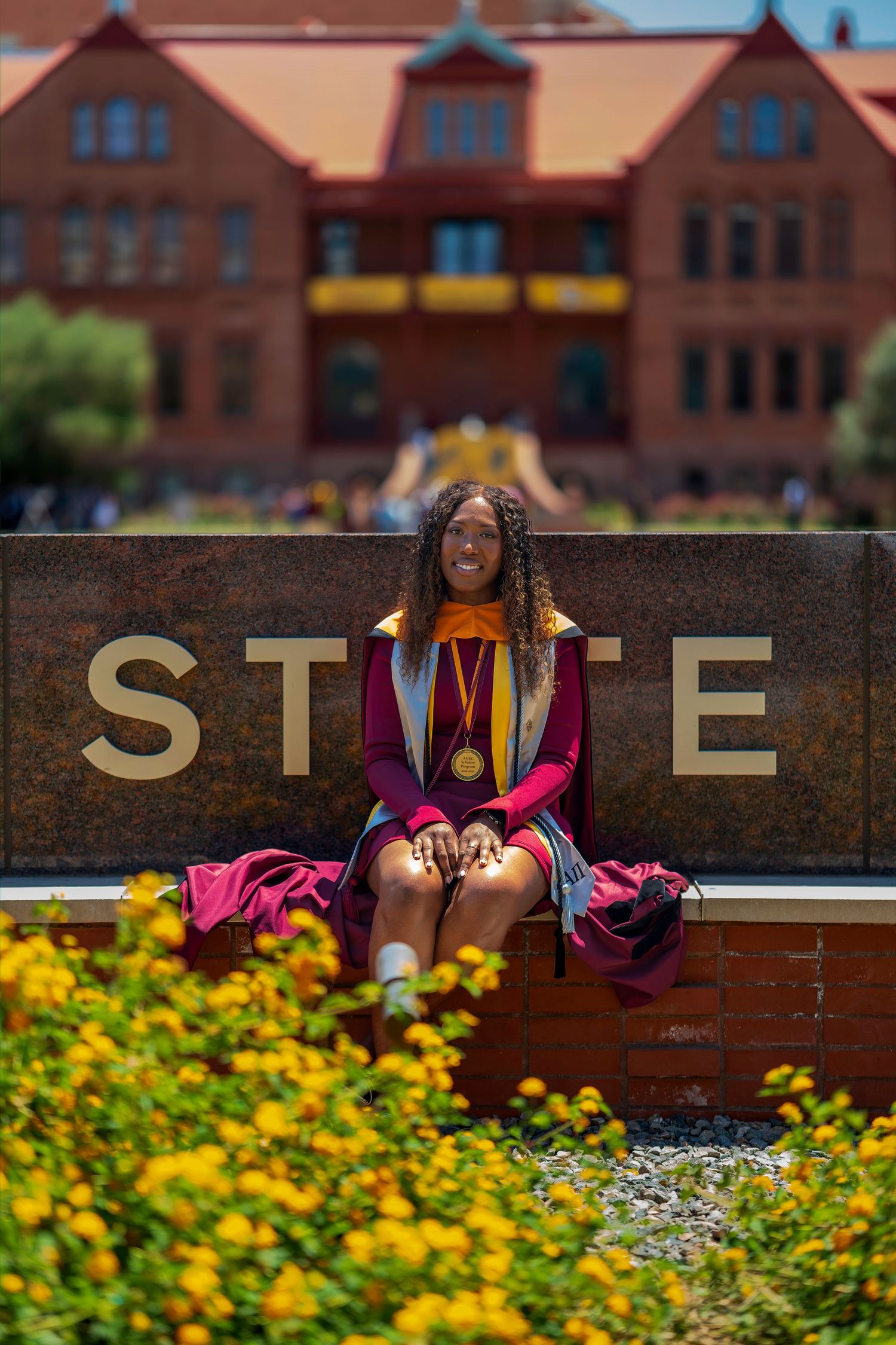 Smiling graduate in maroon regalia sits on STATE sign surrounded by yellow flowers on sunny campus.