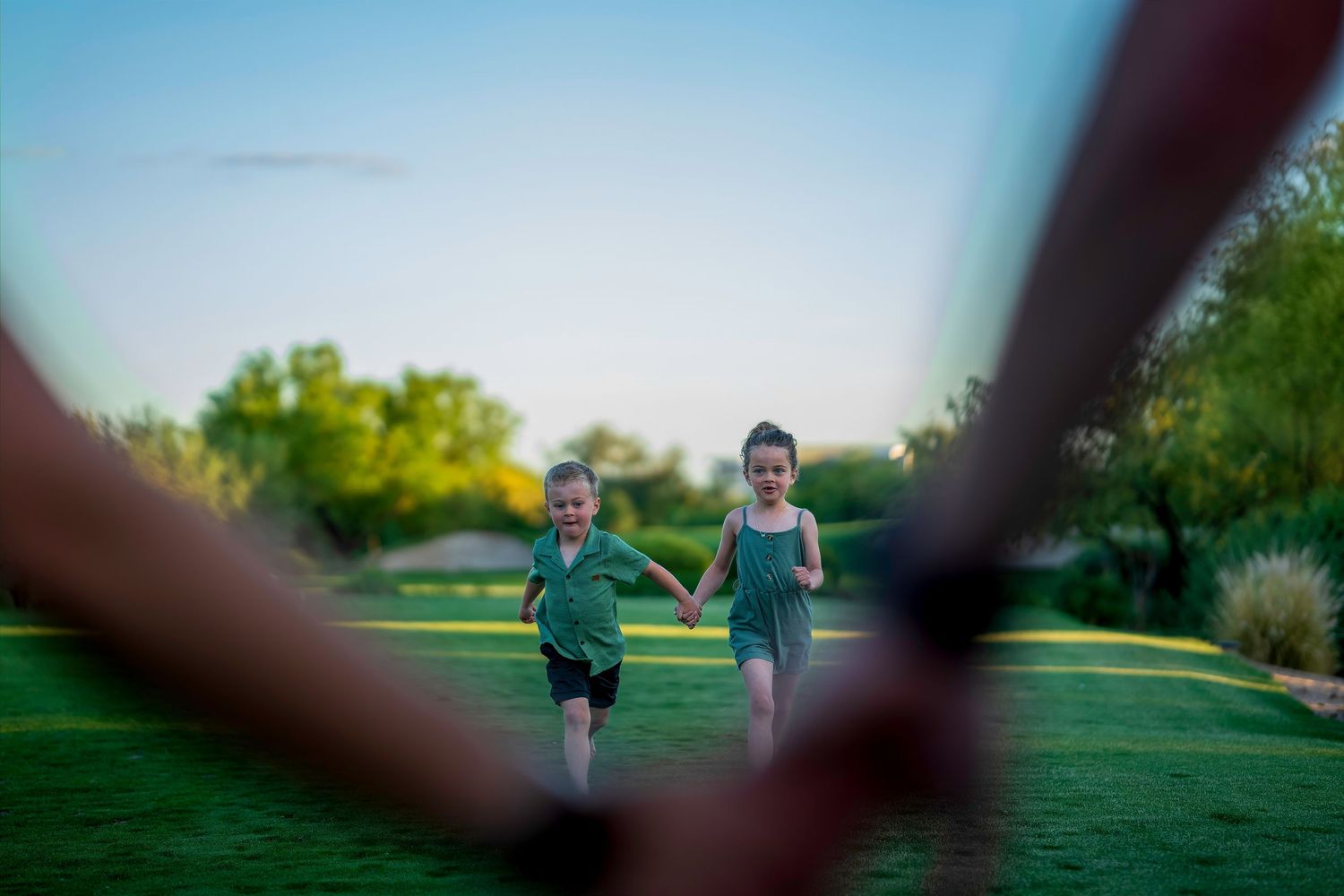 Two young children running hand-in-hand through a green park at dusk.