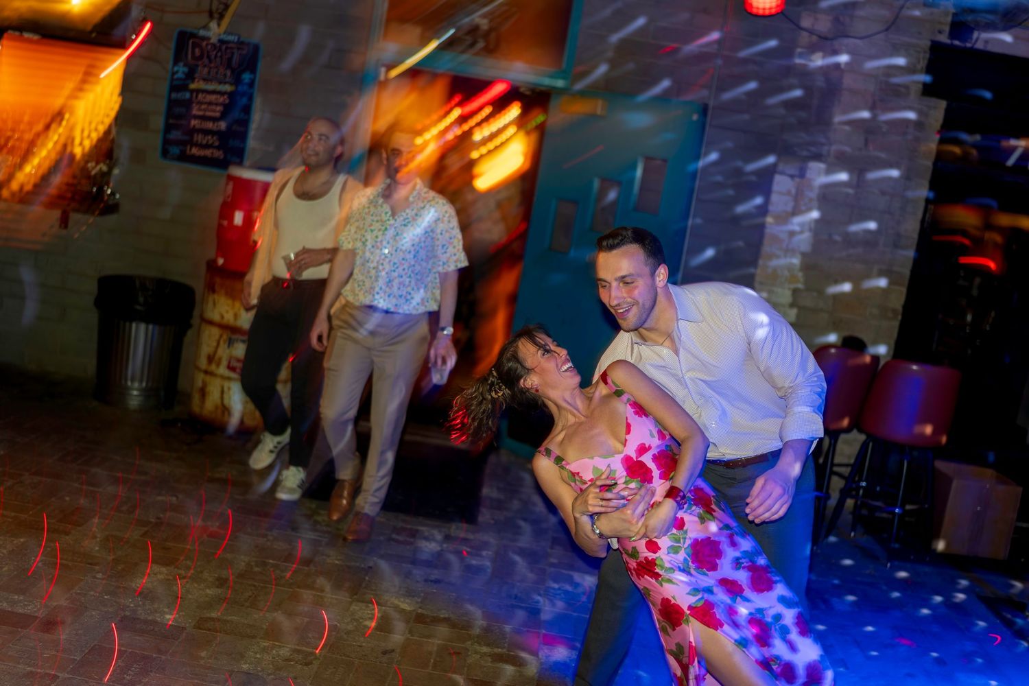 Couple dancing romantically on a colorful dance floor, with another man watching in a dimly lit nightclub.