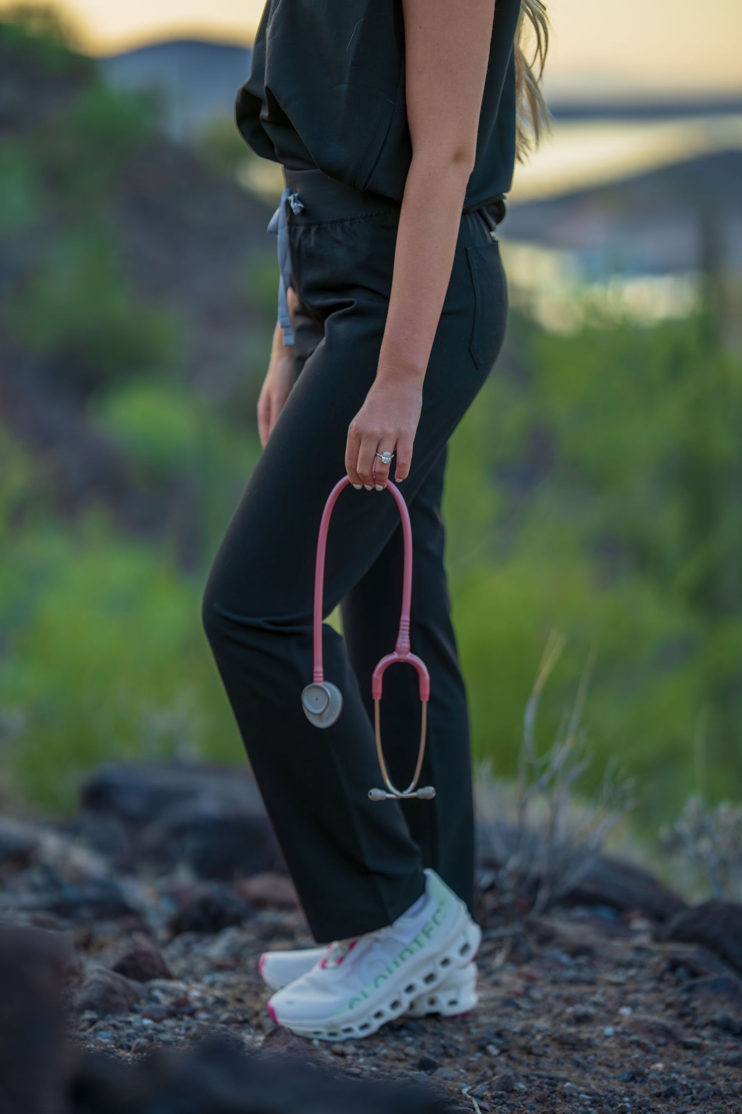 Close-up of pink stethoscope and medical scrubs against desert landscape at dusk.