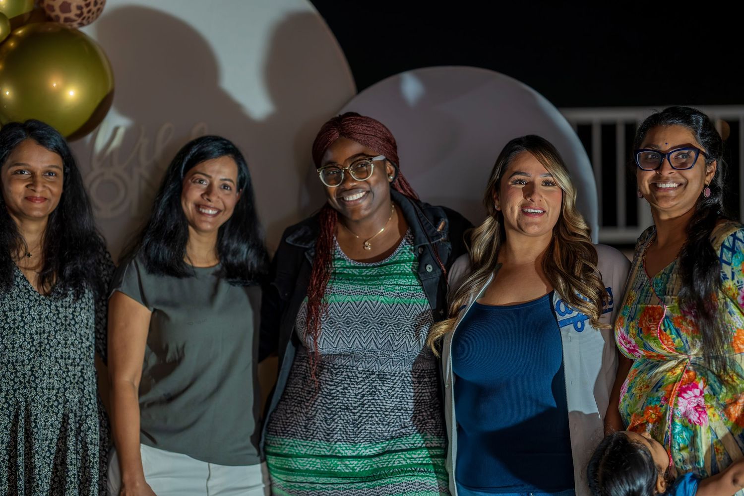 A group of friends smile together at a party against a white wall with dramatic lighting.