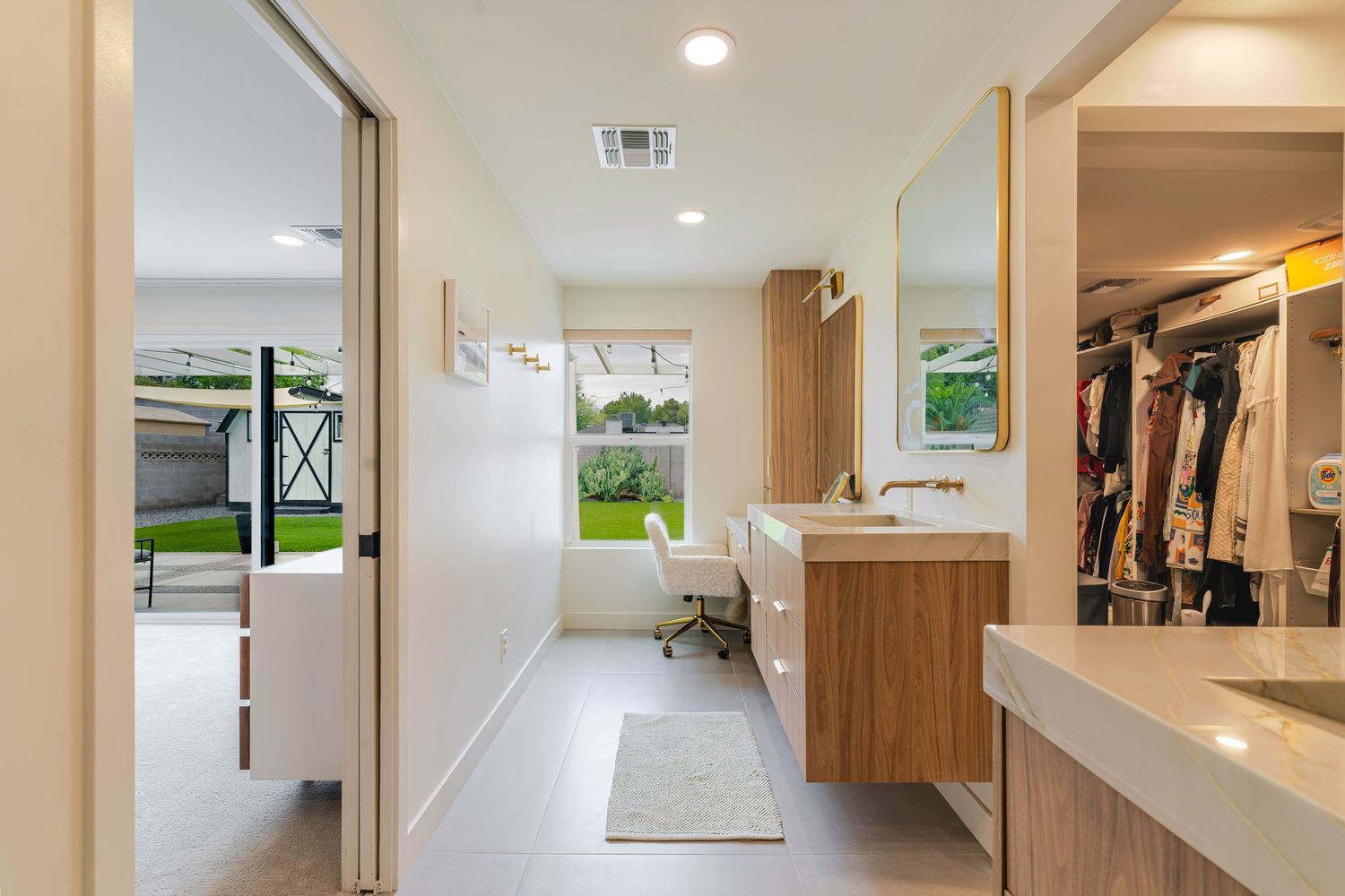 Bright walk-in closet and vanity area with wood cabinetry, white countertop, and view to lush backyard.