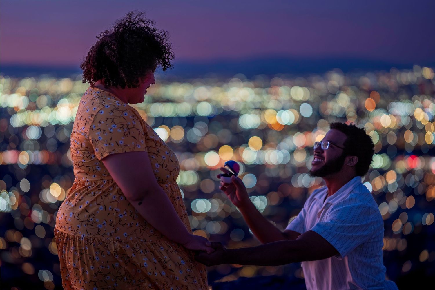 Man kneeling on one knee proposing to woman with city lights glowing in background at night.