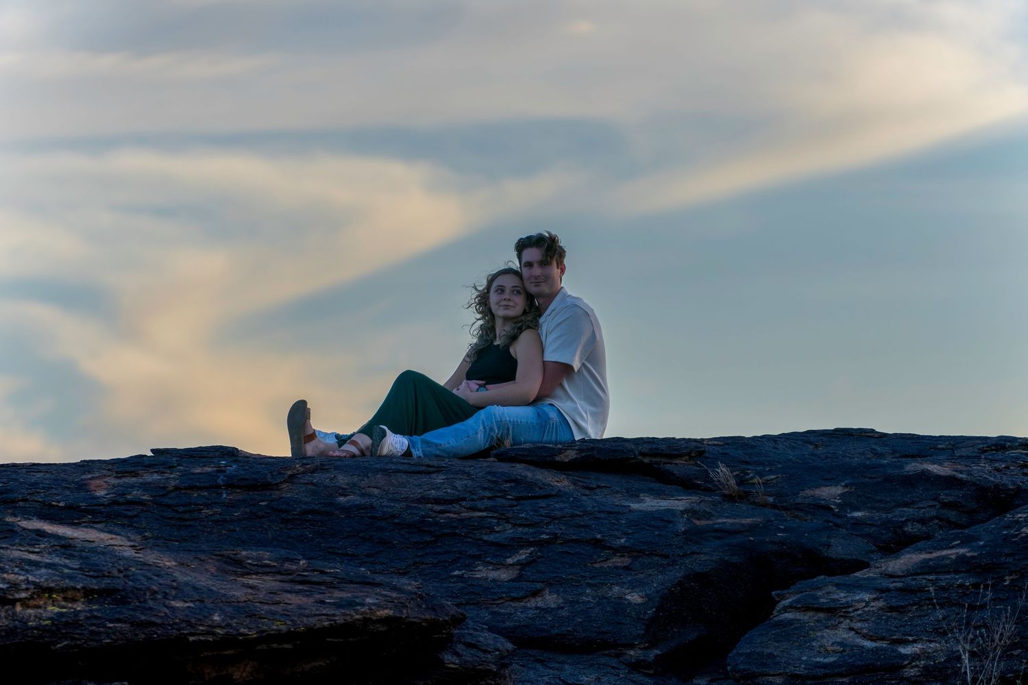 Couple sitting together on rocky outcrop at sunset with dramatic cloudy sky backdrop.