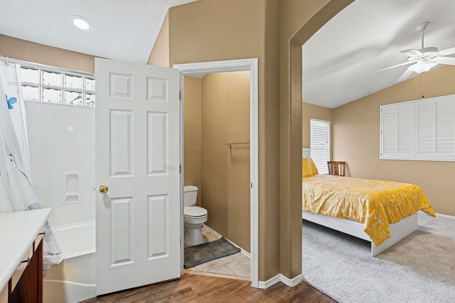 Bright bedroom with vaulted ceiling, yellow bedding, ceiling fan, and an en-suite bathroom with toilet and walk-in shower.