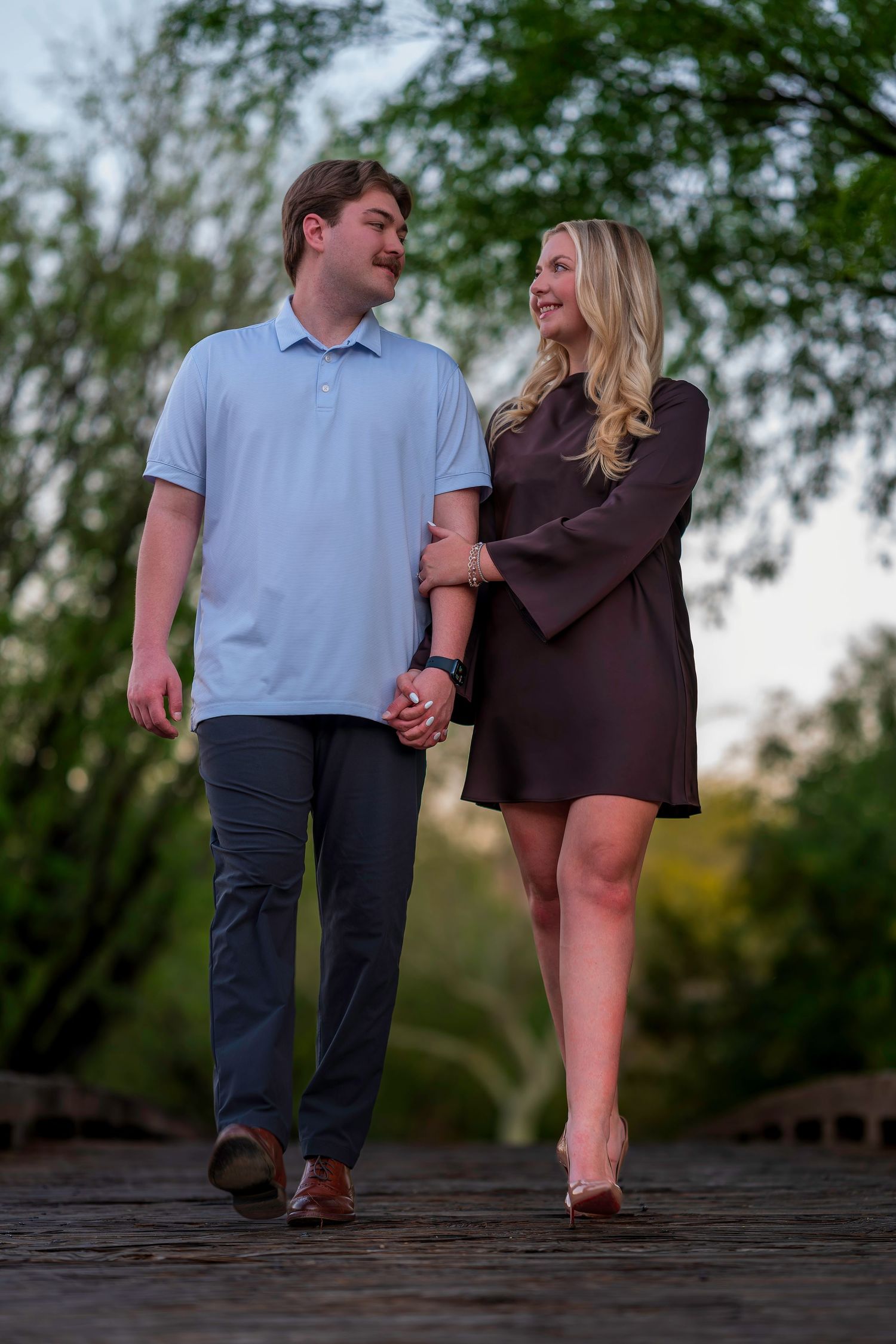 Happy couple holding hands and walking together on a wooden bridge surrounded by green trees.