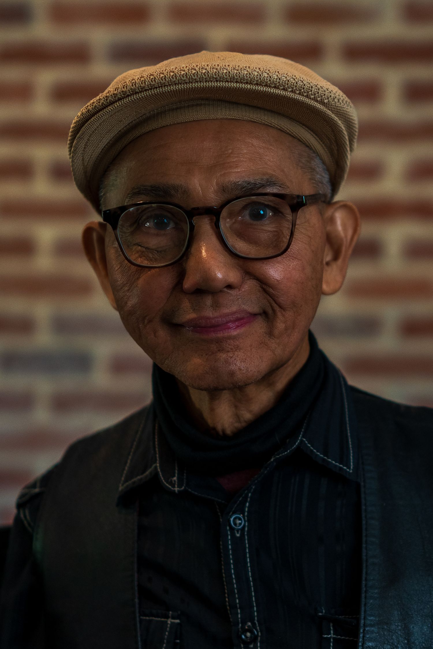 Portrait of someone wearing glasses and a flat cap against a brick wall.