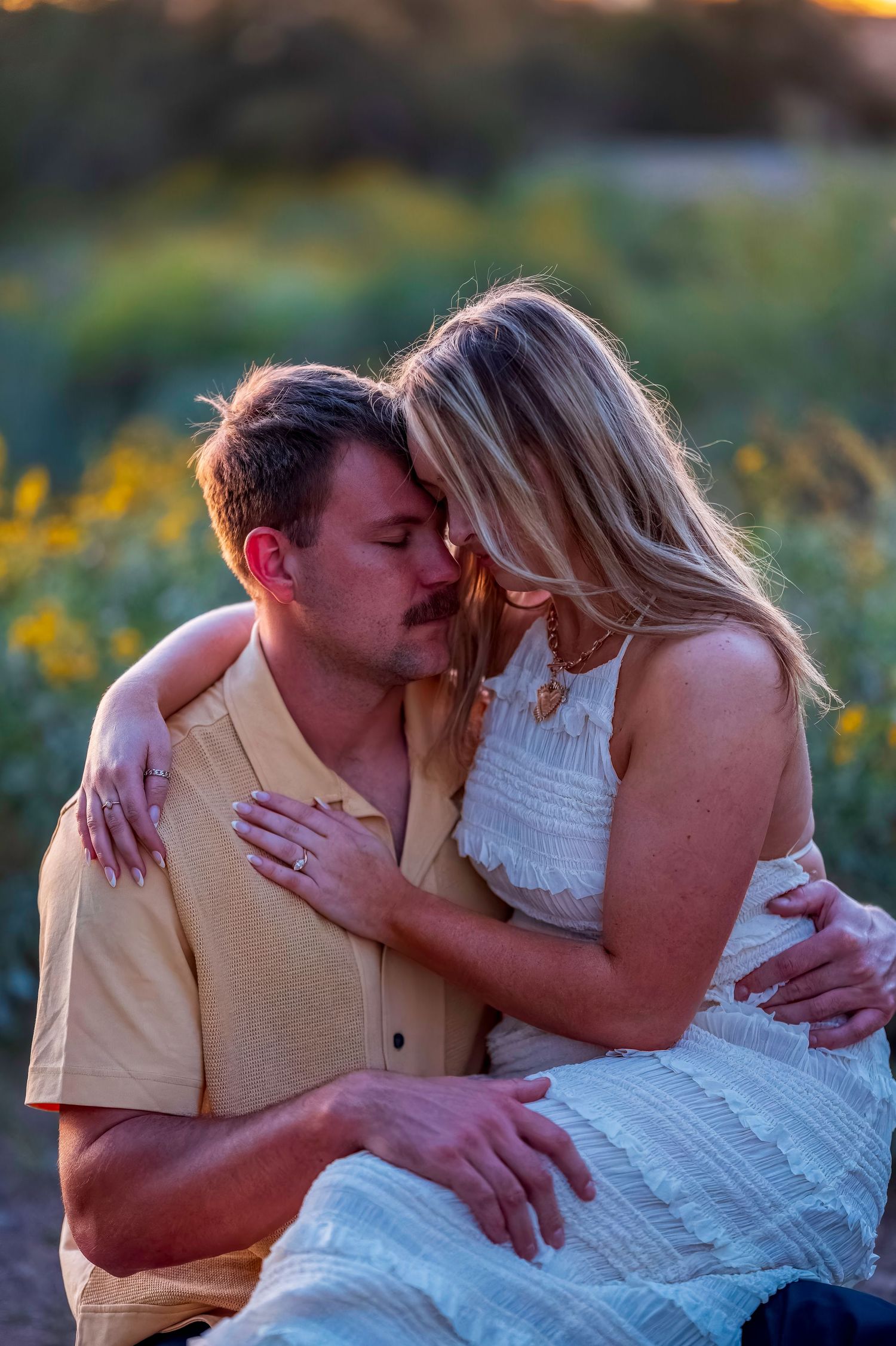 Couple sharing a romantic moment at golden hour in a wildflower field, woman wearing engagement ring in white dress.