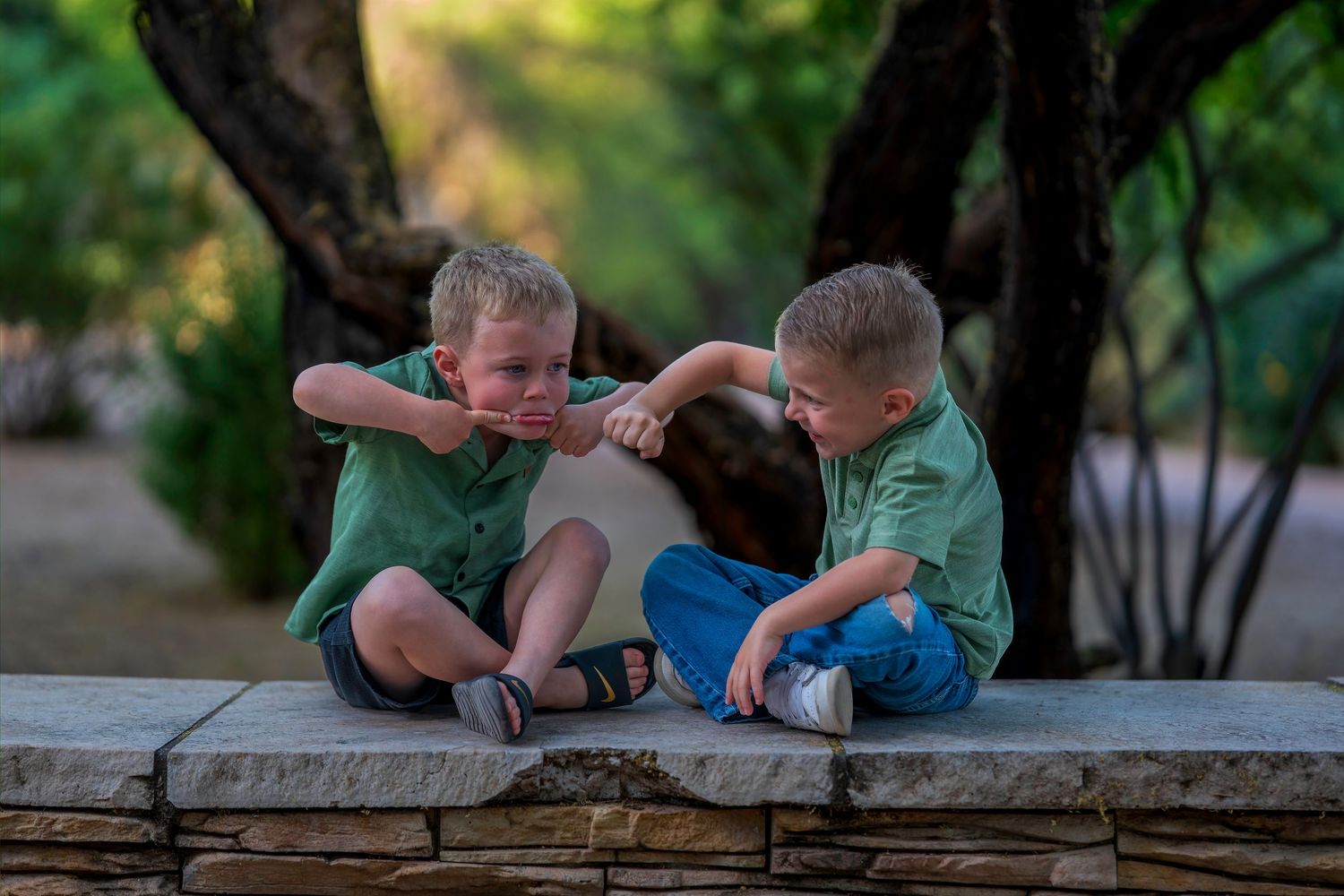 Three young boys in green shirts play together on a stone wall outdoors, surrounded by lush green trees.