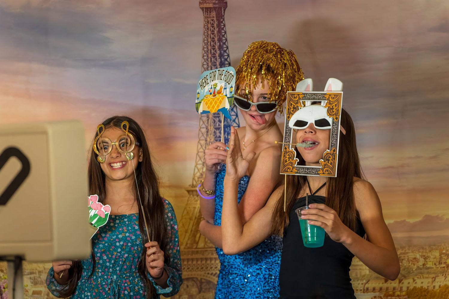 Three girls pose with fun photo booth props including glasses and a frame in front of an Eiffel Tower backdrop.
