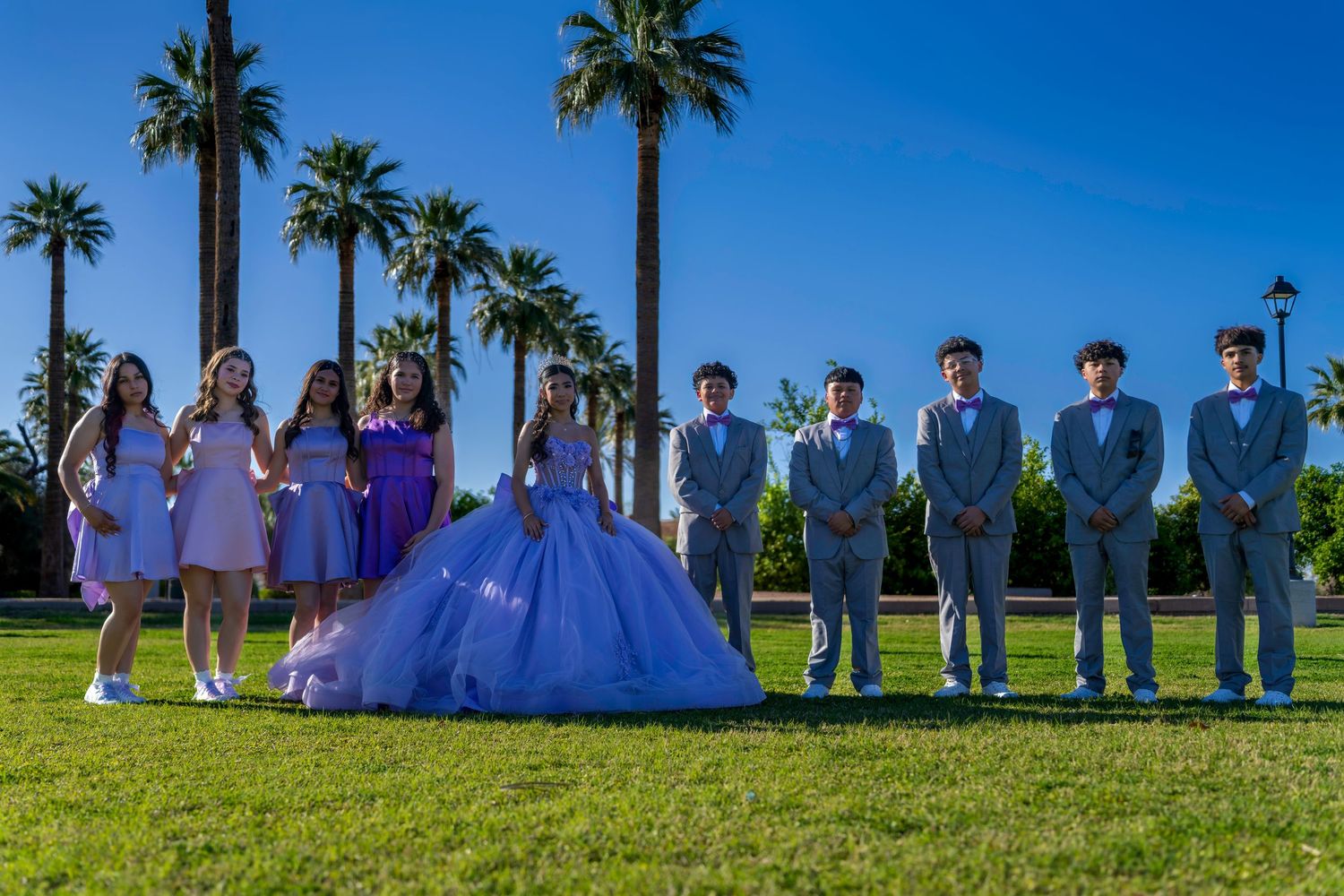 A quinceañera celebration with girls in purple dresses and boys in gray suits posing outdoors near palm trees under a blue sky.