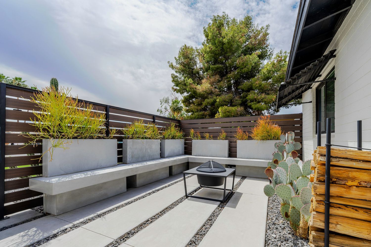 Contemporary outdoor patio with concrete bench, fire pit, wooden privacy fence, and raised planter boxes with greenery.