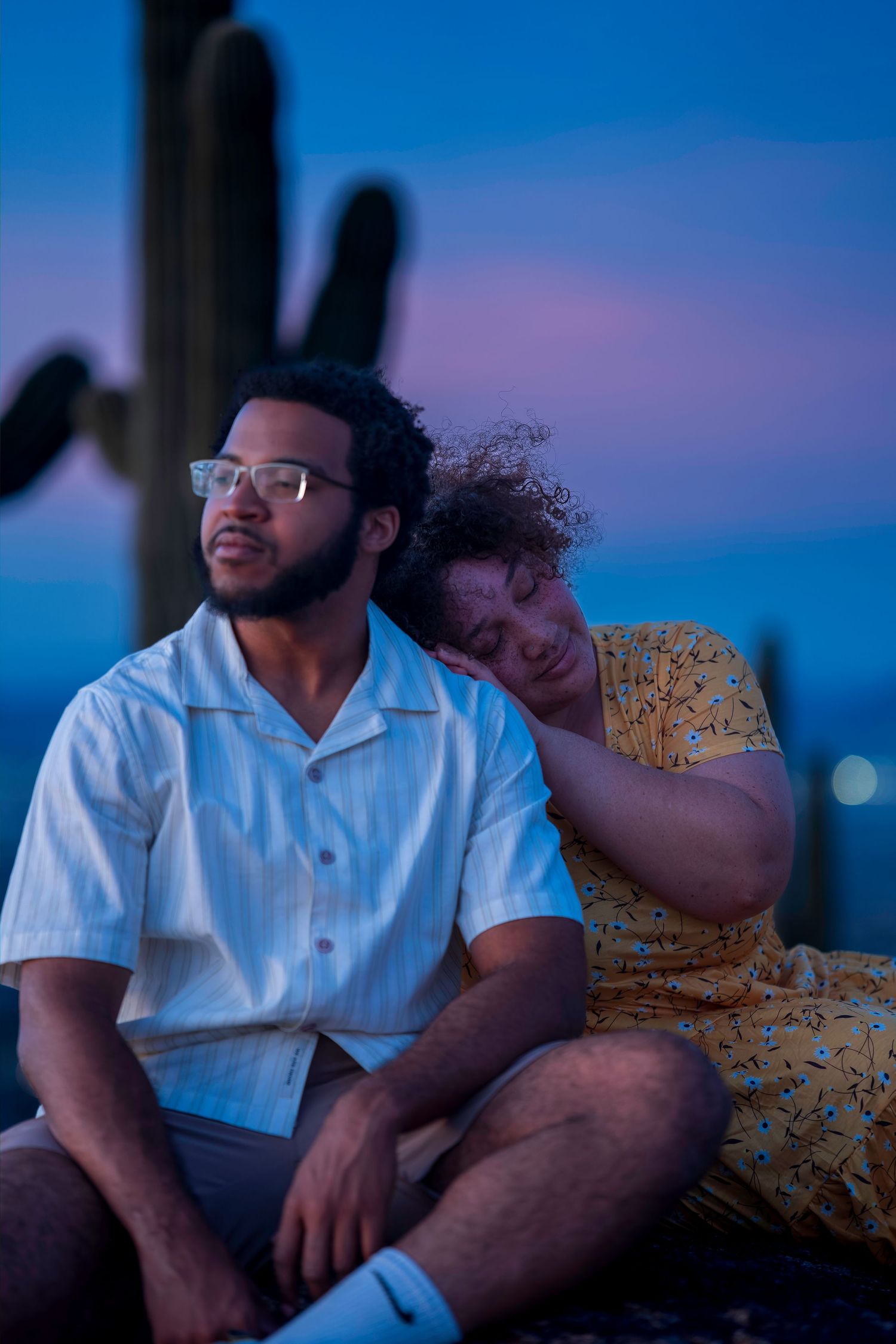 Couple sitting together at dusk near saguaro cacti, woman resting head on mans shoulder under purple sky.