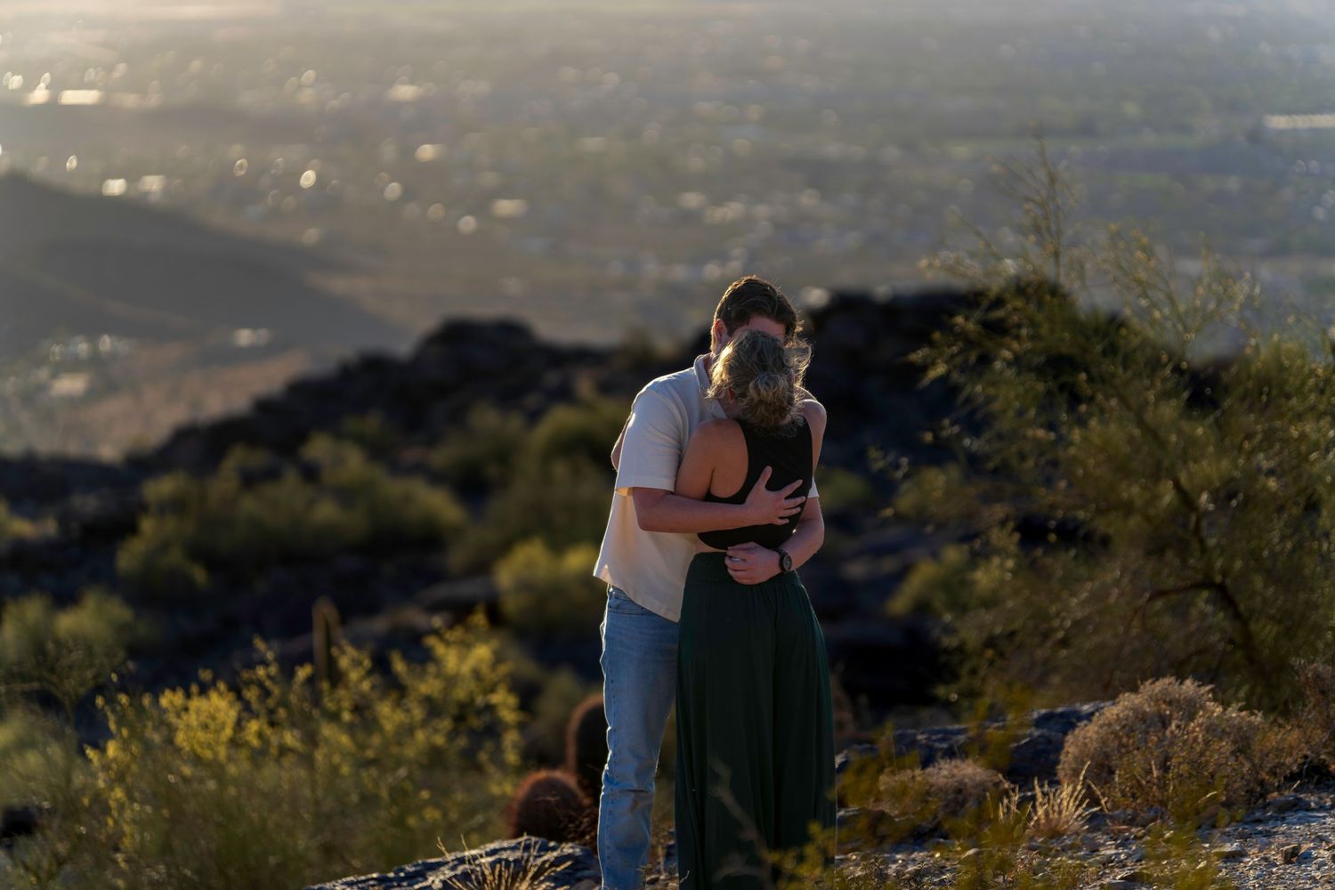Couple embracing on a rocky hilltop at sunset with a scenic valley cityscape in the background.