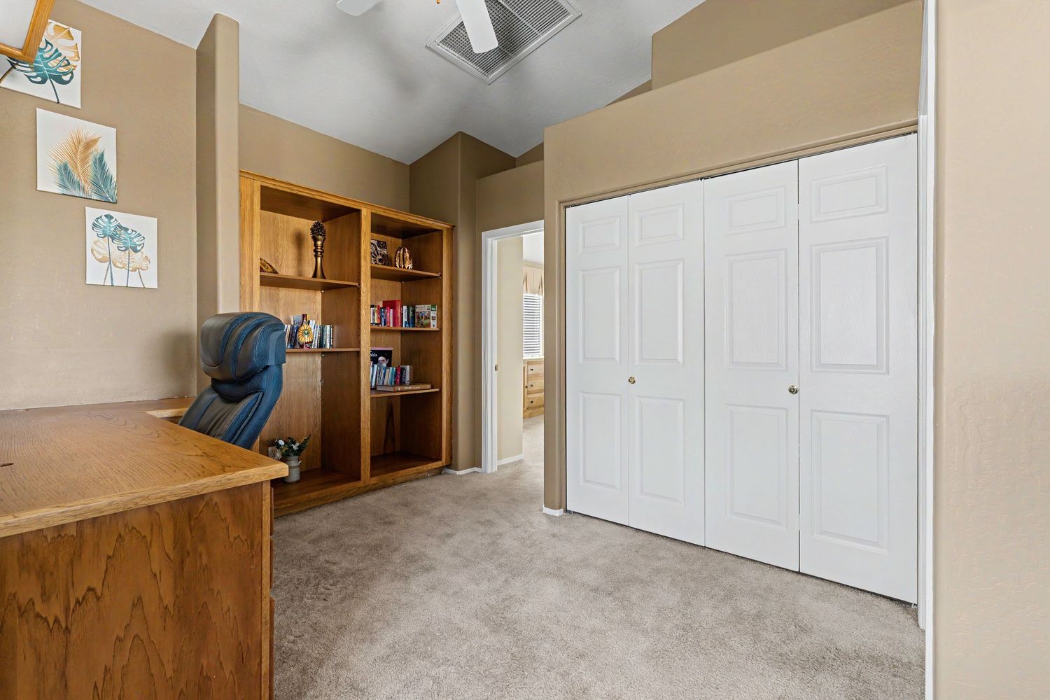 Home office with wood desk, bookshelf, ceiling fan, plush carpet, and white double closet doors in a neutral-toned room.