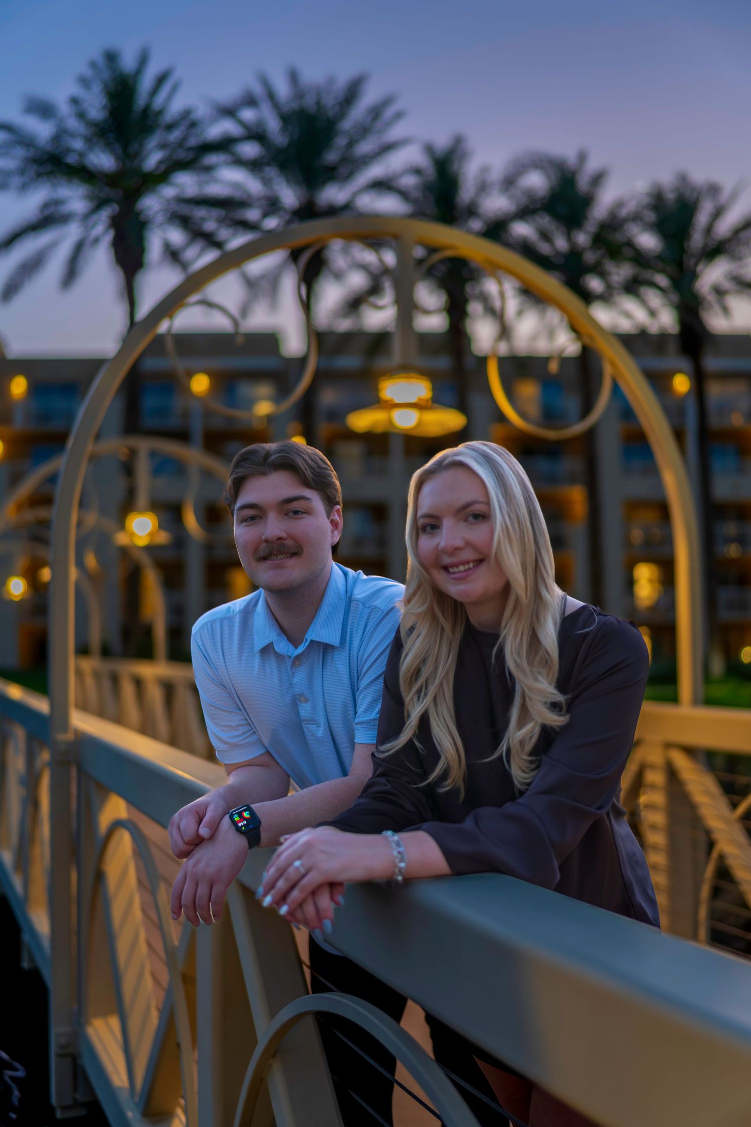 Couple smiling on a decorative bridge at dusk with palm trees and hotel lights in background.
