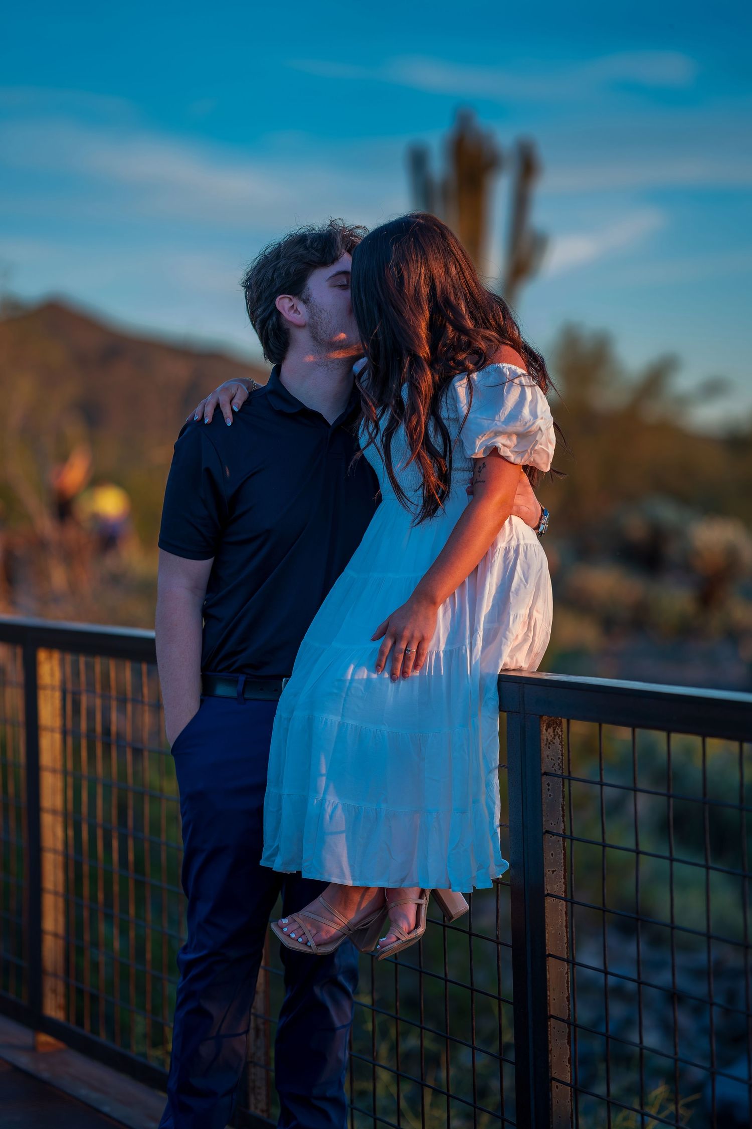 Couple in contrasting black and white outfits embrace by a railing during sunset in a desert setting.