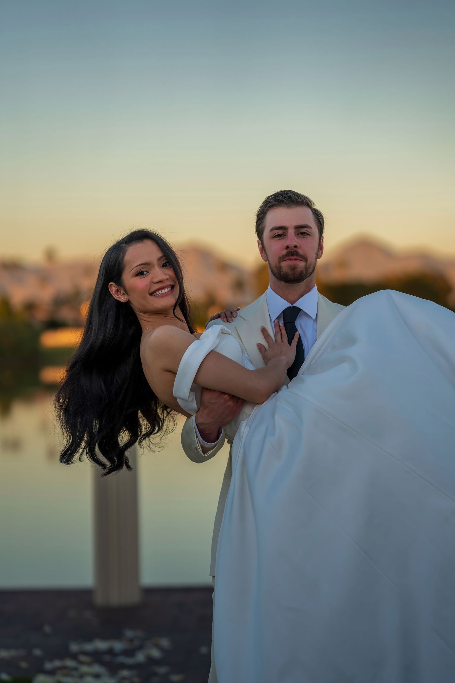 Playful wedding couple dancing at waterfront during sunset.