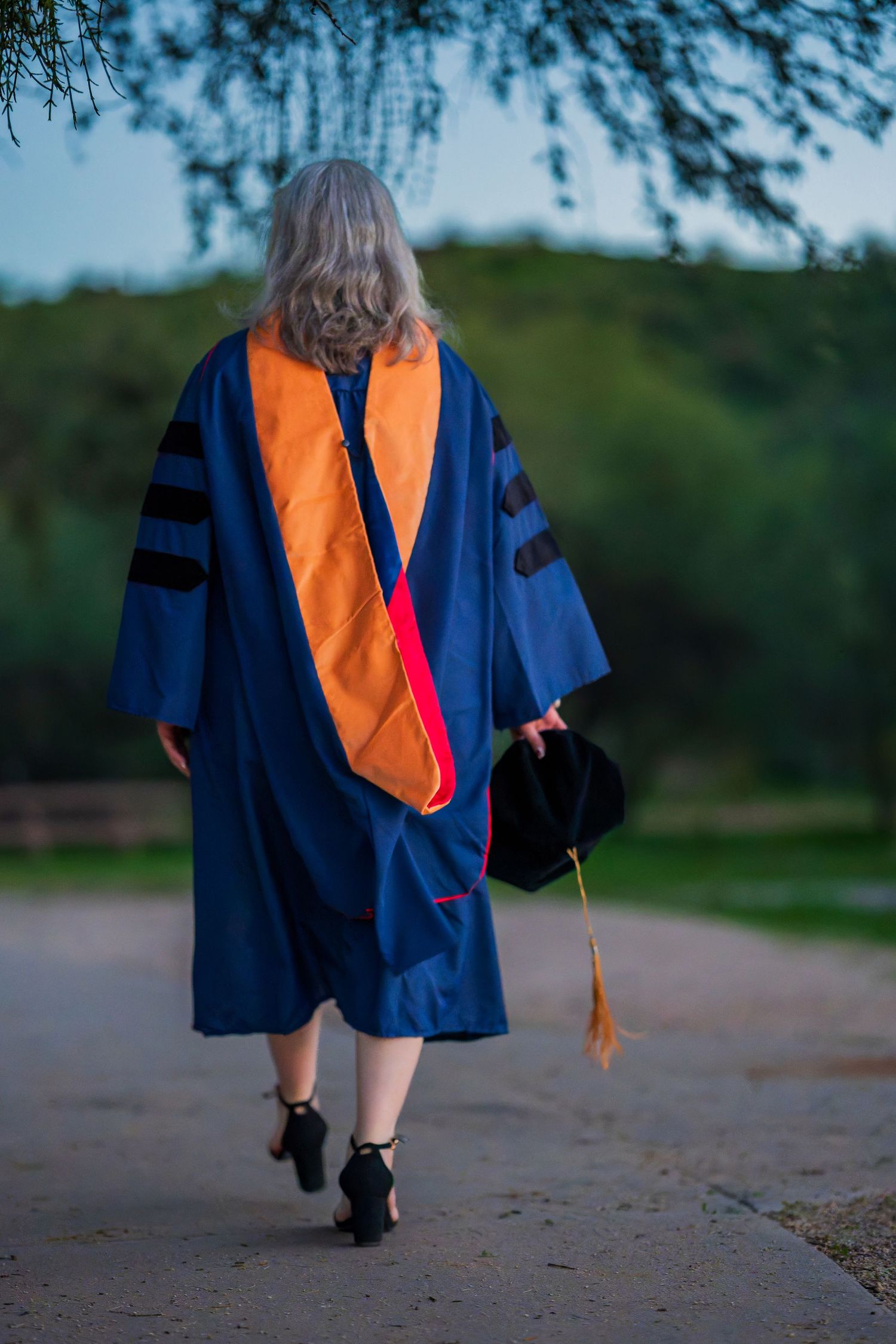 Graduate in blue and orange academic regalia walking on a path during commencement.