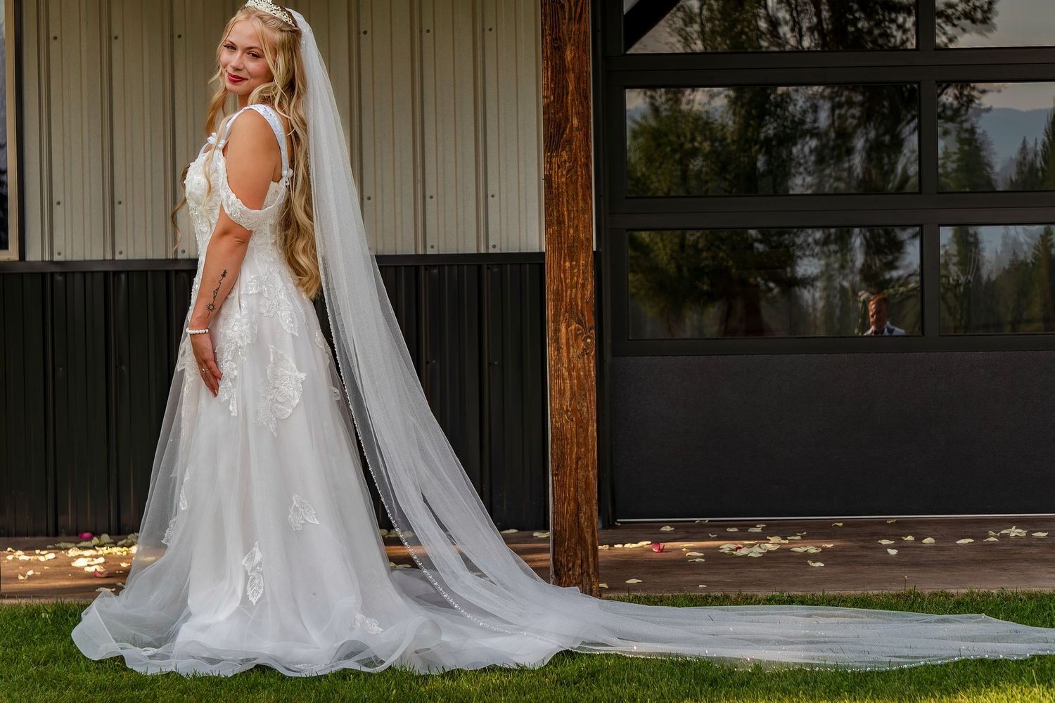 A bride in a flowing white lace gown with long train poses against a rustic building backdrop.
