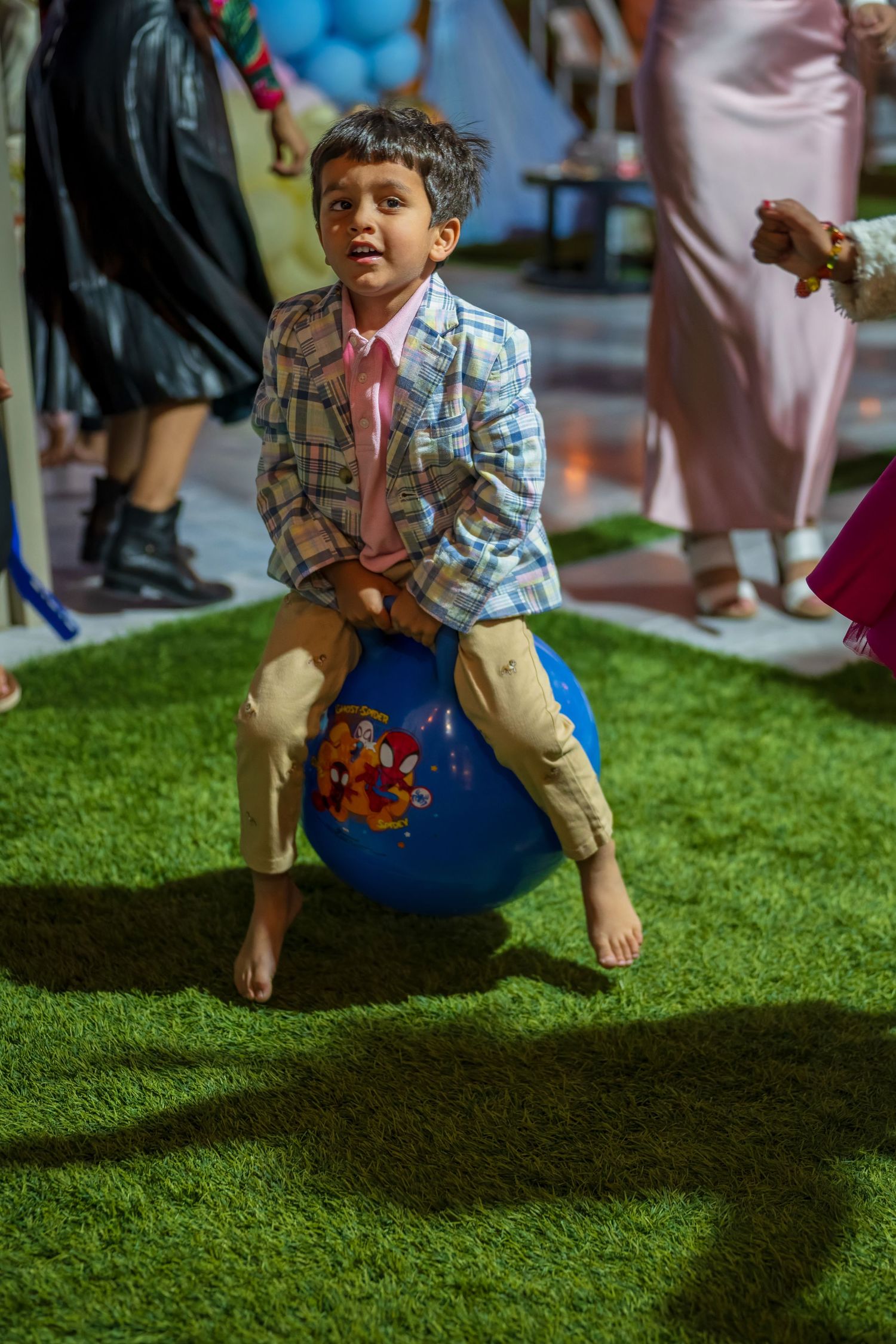 A child plays on artificial grass during an indoor event with blue balloons in the background.