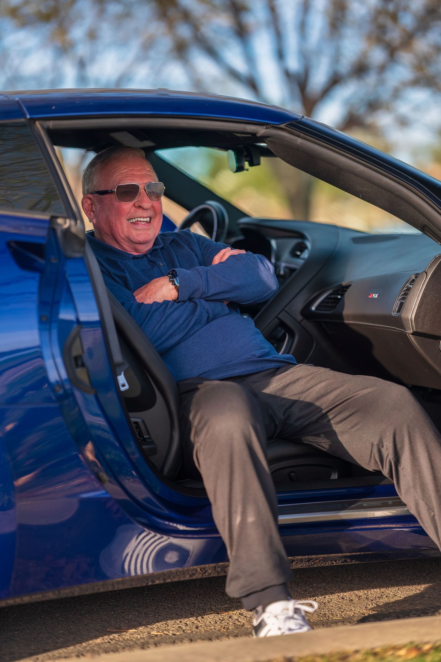 Smiling senior man in sunglasses sitting in blue sports car with door open, arms crossed, wearing navy jacket.