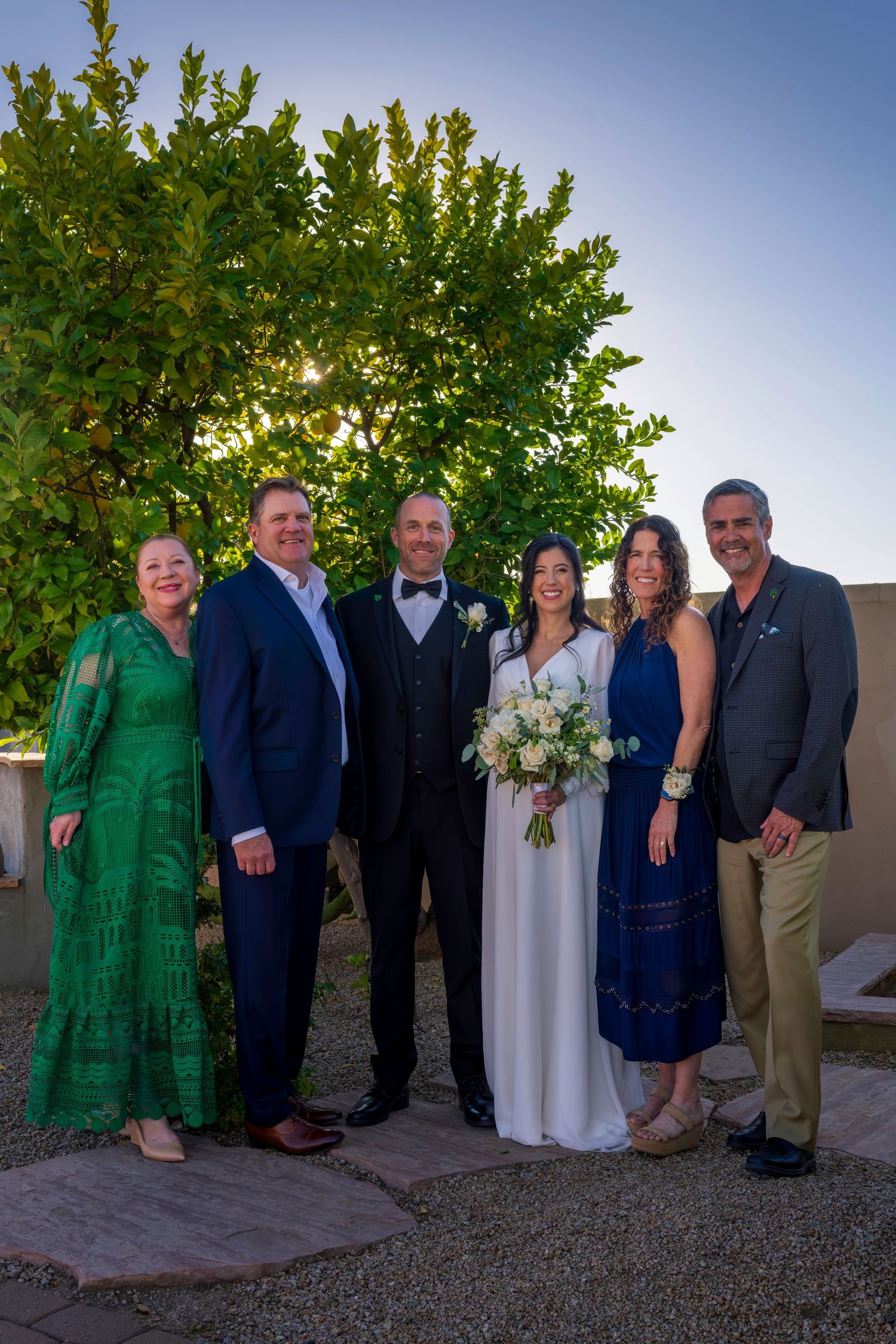 Wedding couple posing with four family members outdoors at dusk, bride holding white floral bouquet.