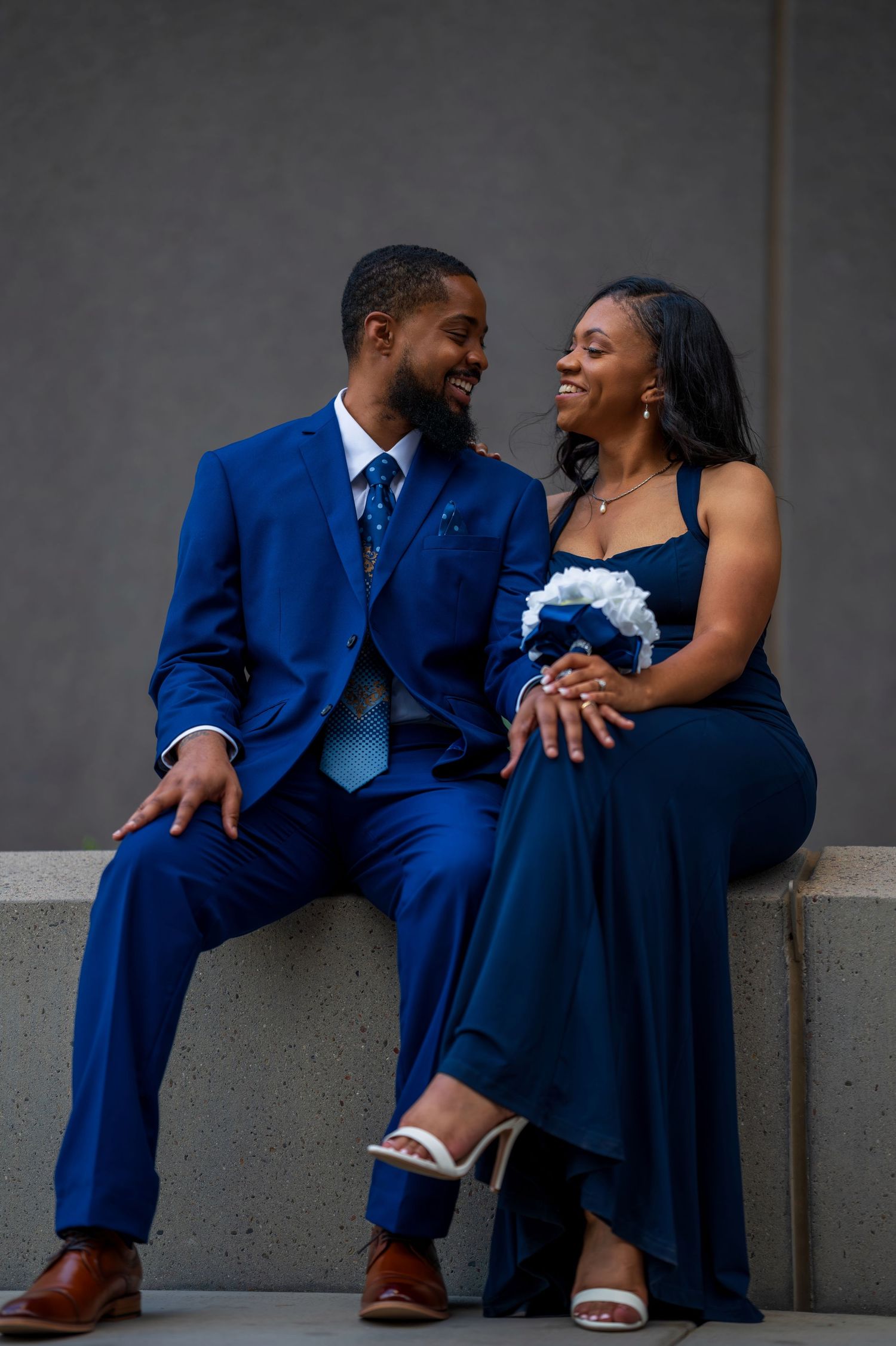 Couple in matching blue formal wear sitting together smiling, man in suit and woman holding bouquet.