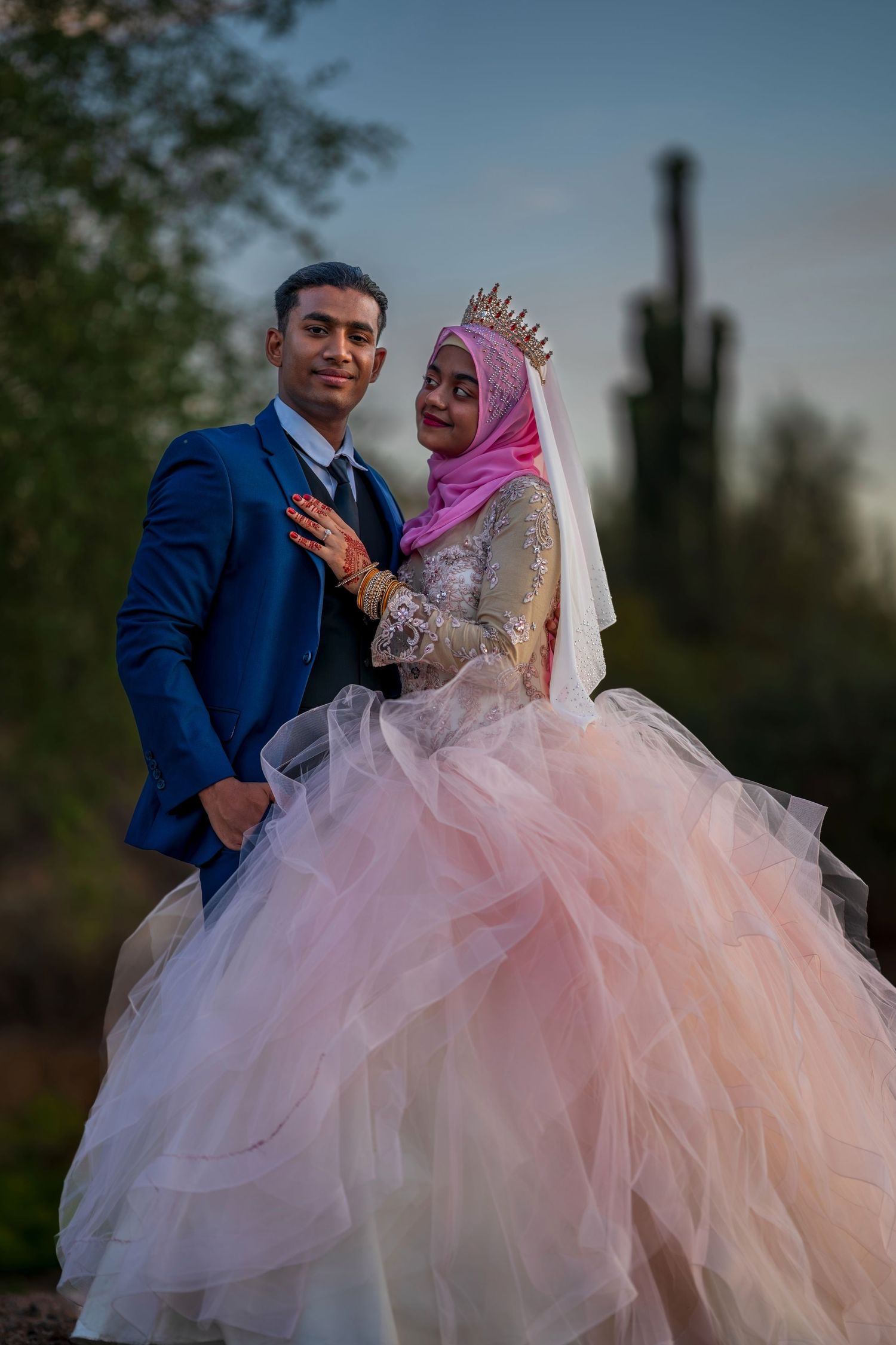 Muslim bride in pink tulle gown and hijab with groom in blue suit posing outdoors at sunset.