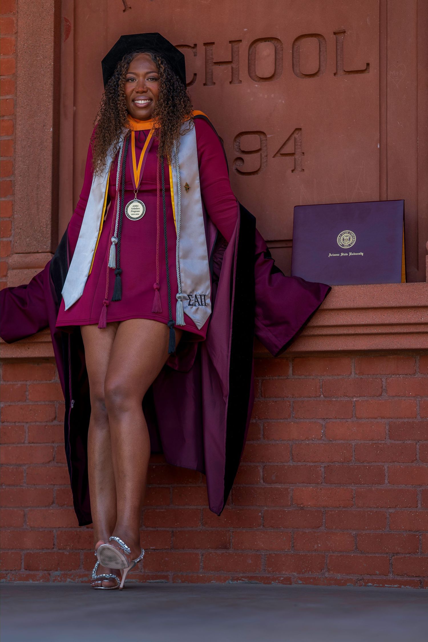 Proud graduate in maroon doctoral gown with medal and diploma at ASU Normal School 1894 building.