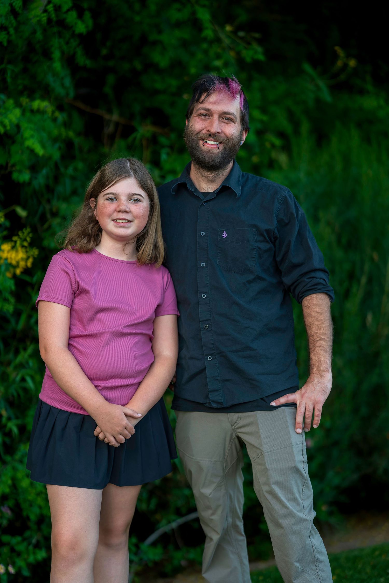 Smiling bearded man and young girl in pink dress posing together outdoors in lush green garden.