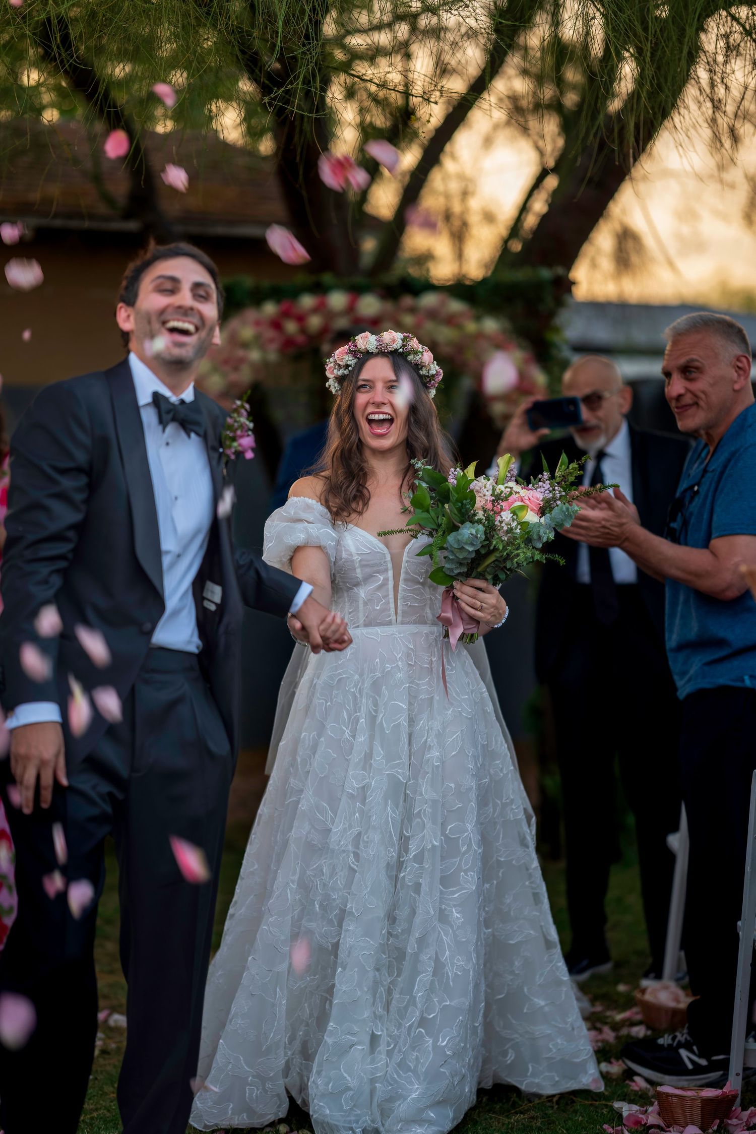 Laughing bride and groom celebrate with rose petal confetti after outdoor wedding ceremony.
