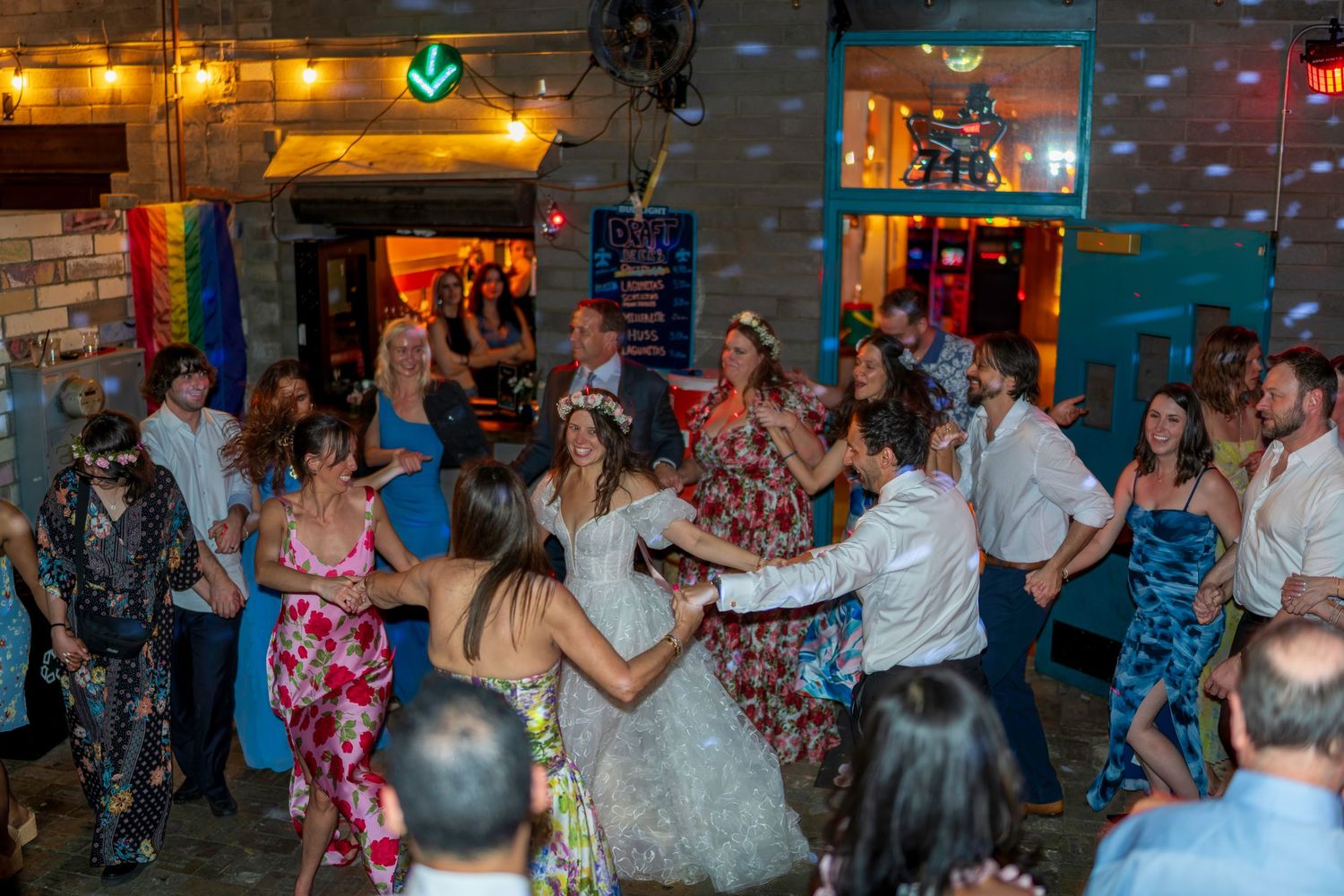 Wedding guests dance joyfully at a lively indoor reception with colorful decor and a rainbow flag in the background.