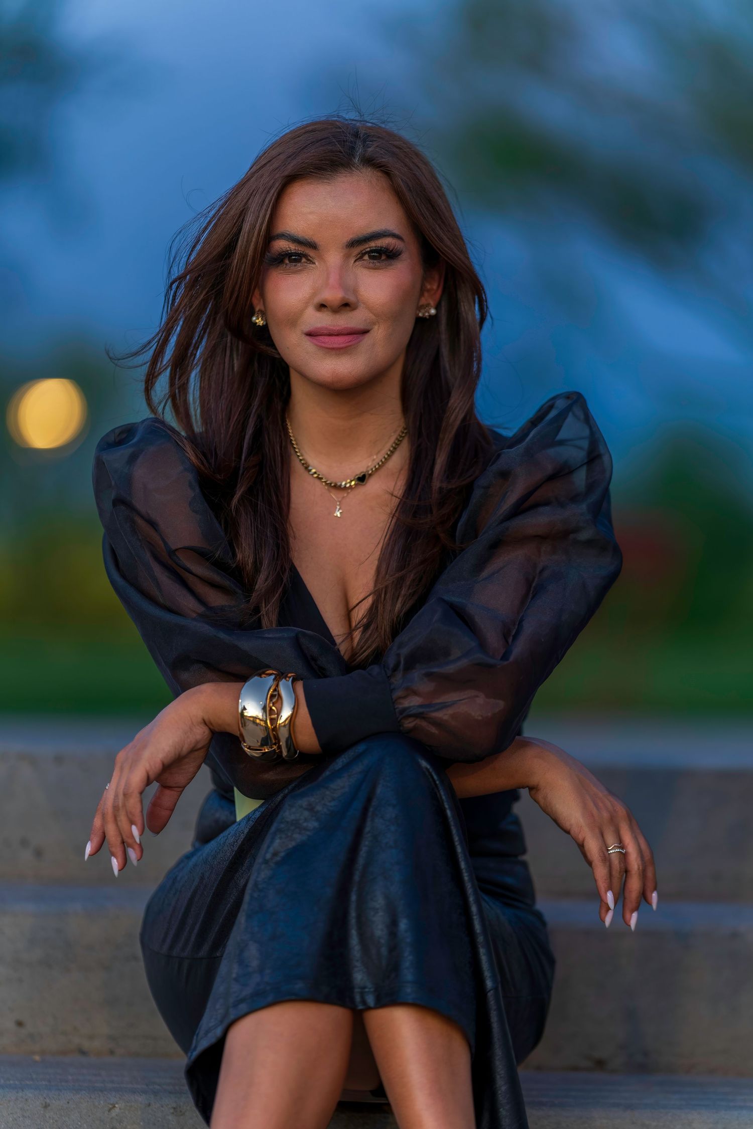 Stylish woman in black sheer dress with gold jewelry sitting on stone steps at twilight.