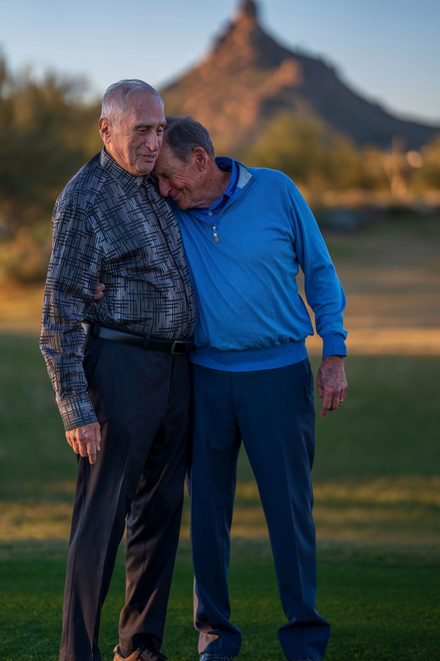 Two people sharing a tender moment while standing on grass with mountain backdrop.