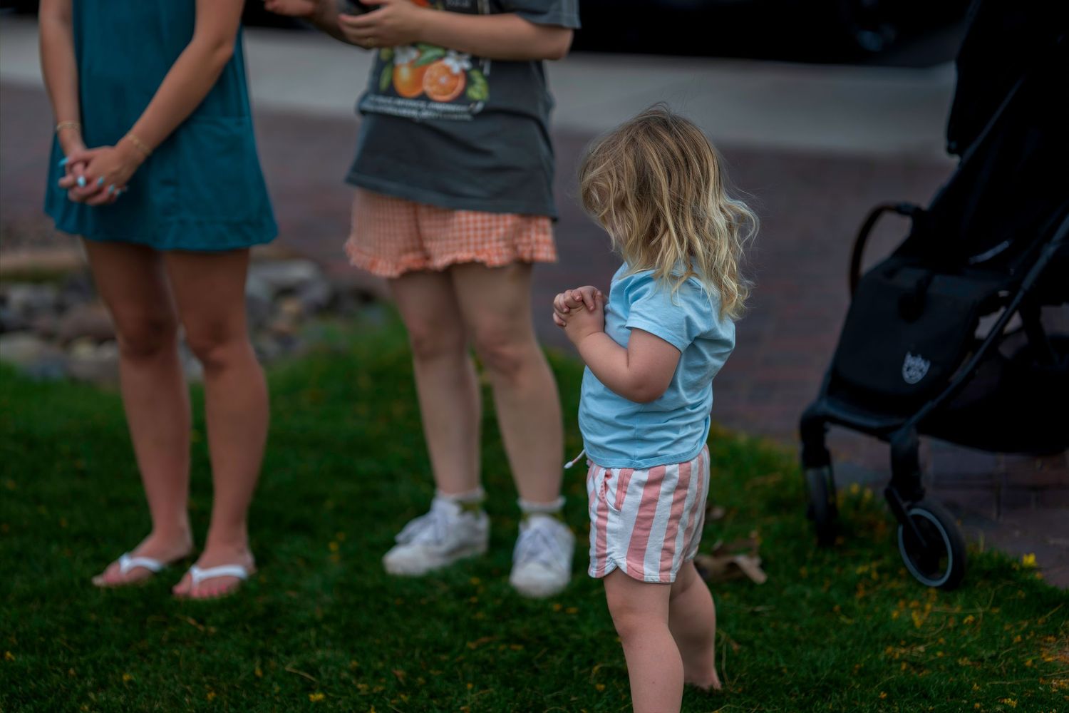 Young toddler girl in blue shirt and striped shorts standing on green grass near adults at outdoor gathering.