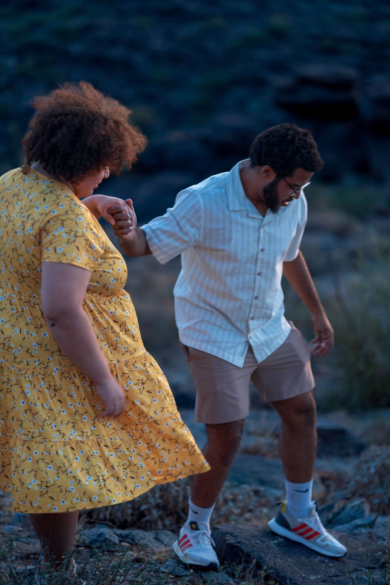 Man helping woman in yellow floral dress navigate rocky terrain during outdoor evening photoshoot.