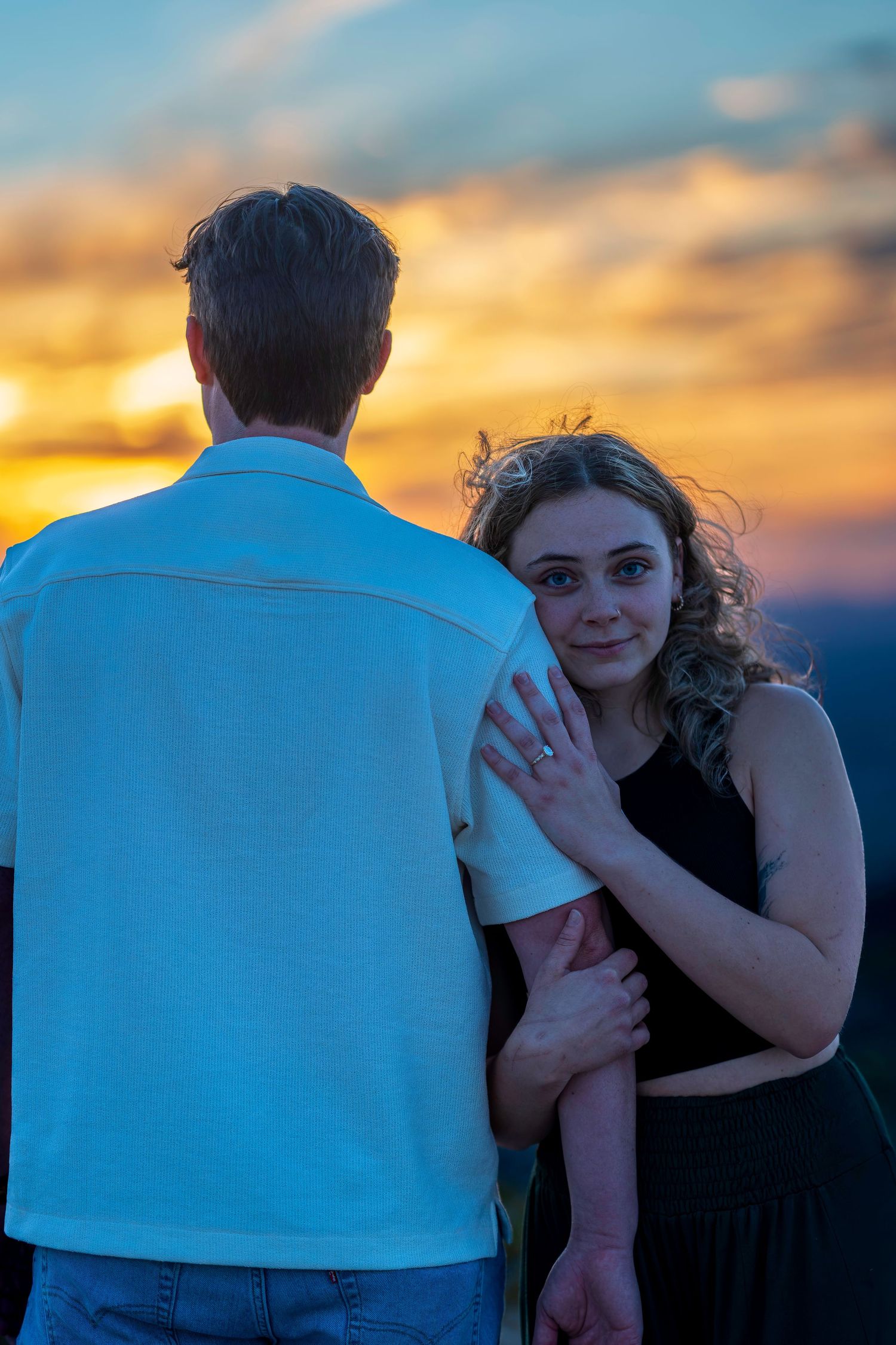 Couple embracing at sunset, woman in black top looks at camera while man in white polo faces away.