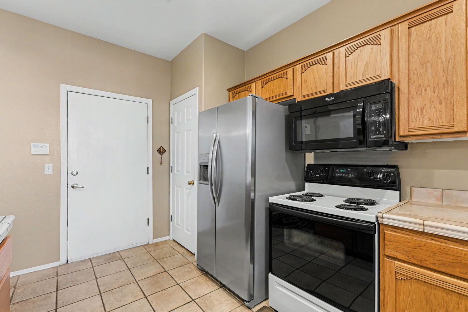 Kitchen with stainless steel refrigerator, white electric range, black microwave, oak cabinets, and light tile flooring.