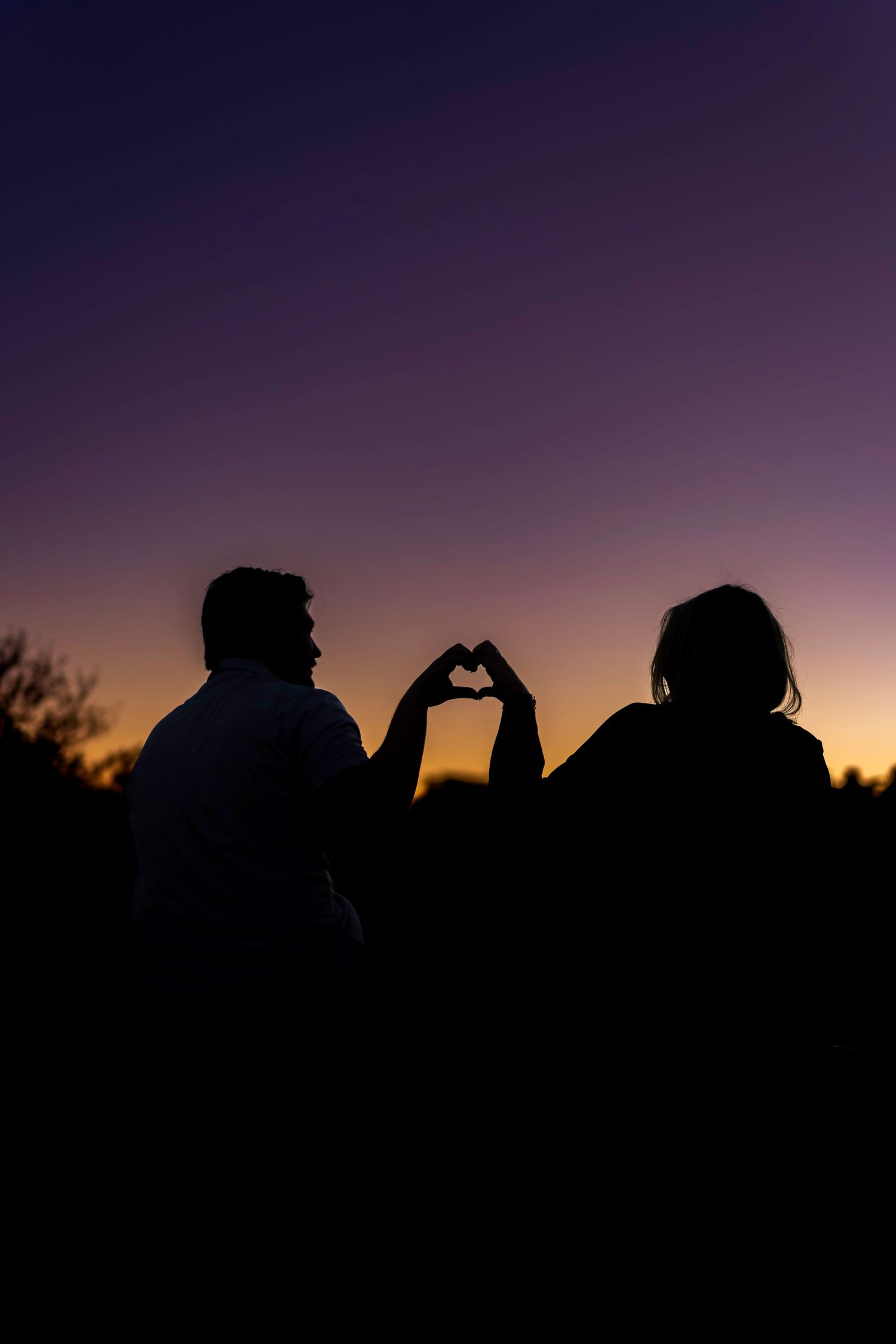 Romantic silhouette of a couple making a heart shape with their hands against a colorful sunset sky.
