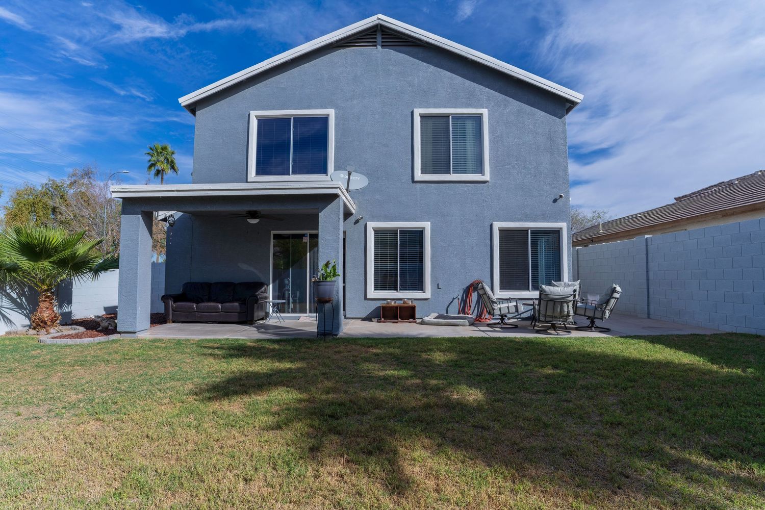Two-story gray stucco home with covered patio, outdoor dining area, green lawn, and palm tree in backyard.