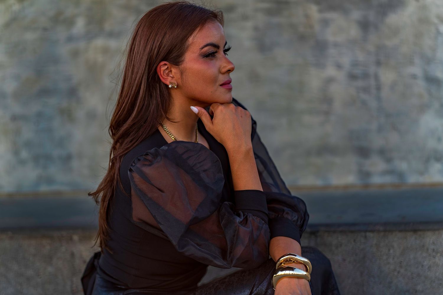 A stylish woman in a sheer black outfit sits outdoors, wearing gold jewelry, gazing thoughtfully to the side.