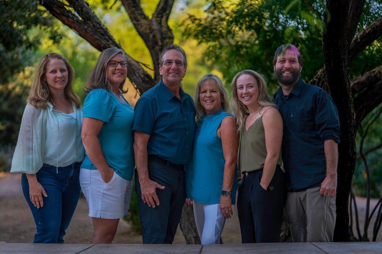 Six smiling family members pose outdoors in coordinating teal and blue outfits surrounded by lush green trees.