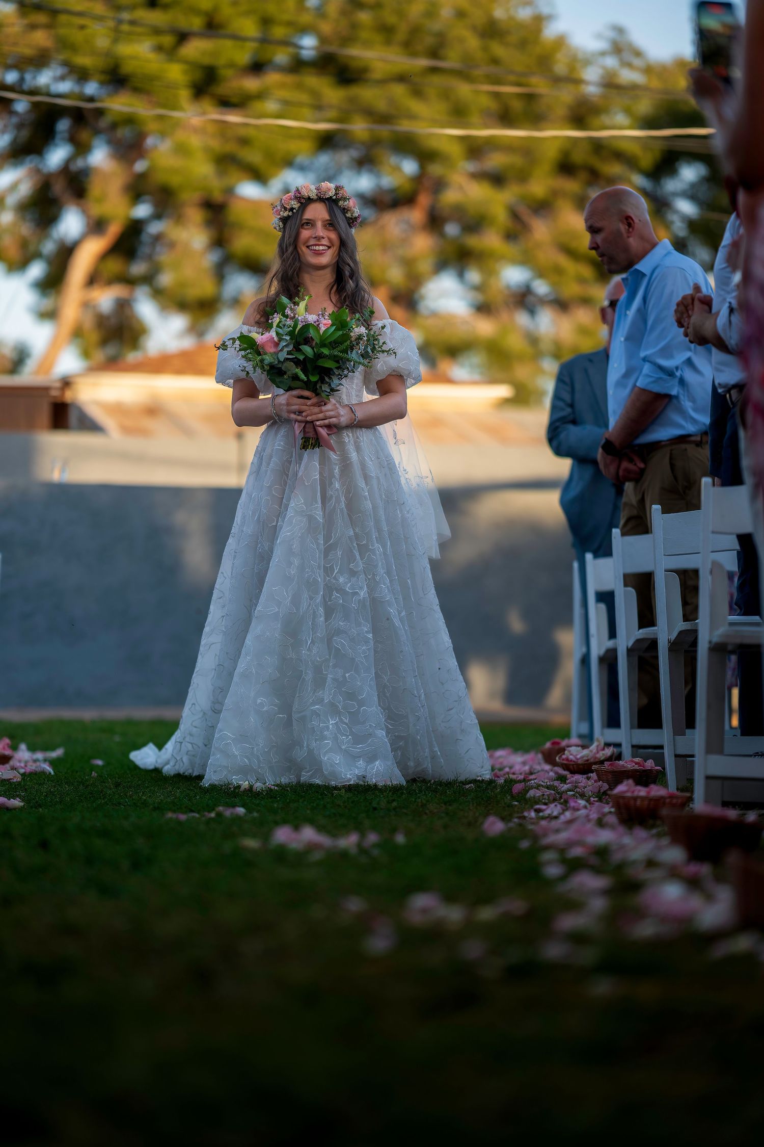 Radiant bride in lace gown and flower crown walking down outdoor wedding aisle smiling.