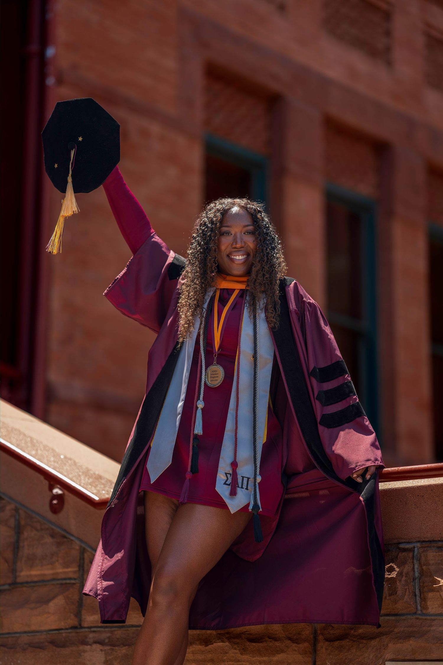 Smiling graduate in maroon doctoral regalia raises cap triumphantly on sunlit steps of brick building.