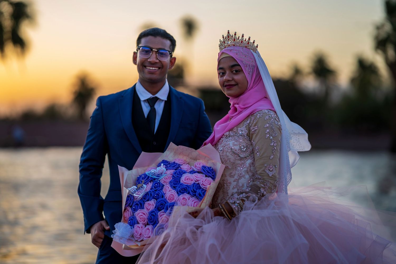 Bride in pink hijab and gown with groom holding bouquet of pink and blue roses at sunset.