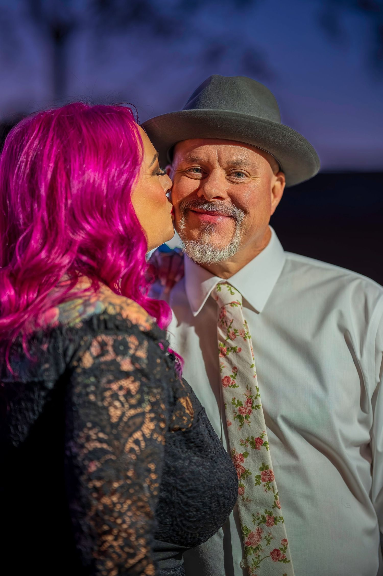 A pink-haired tattooed woman kisses a smiling man in a gray fedora and floral tie on the cheek at dusk.