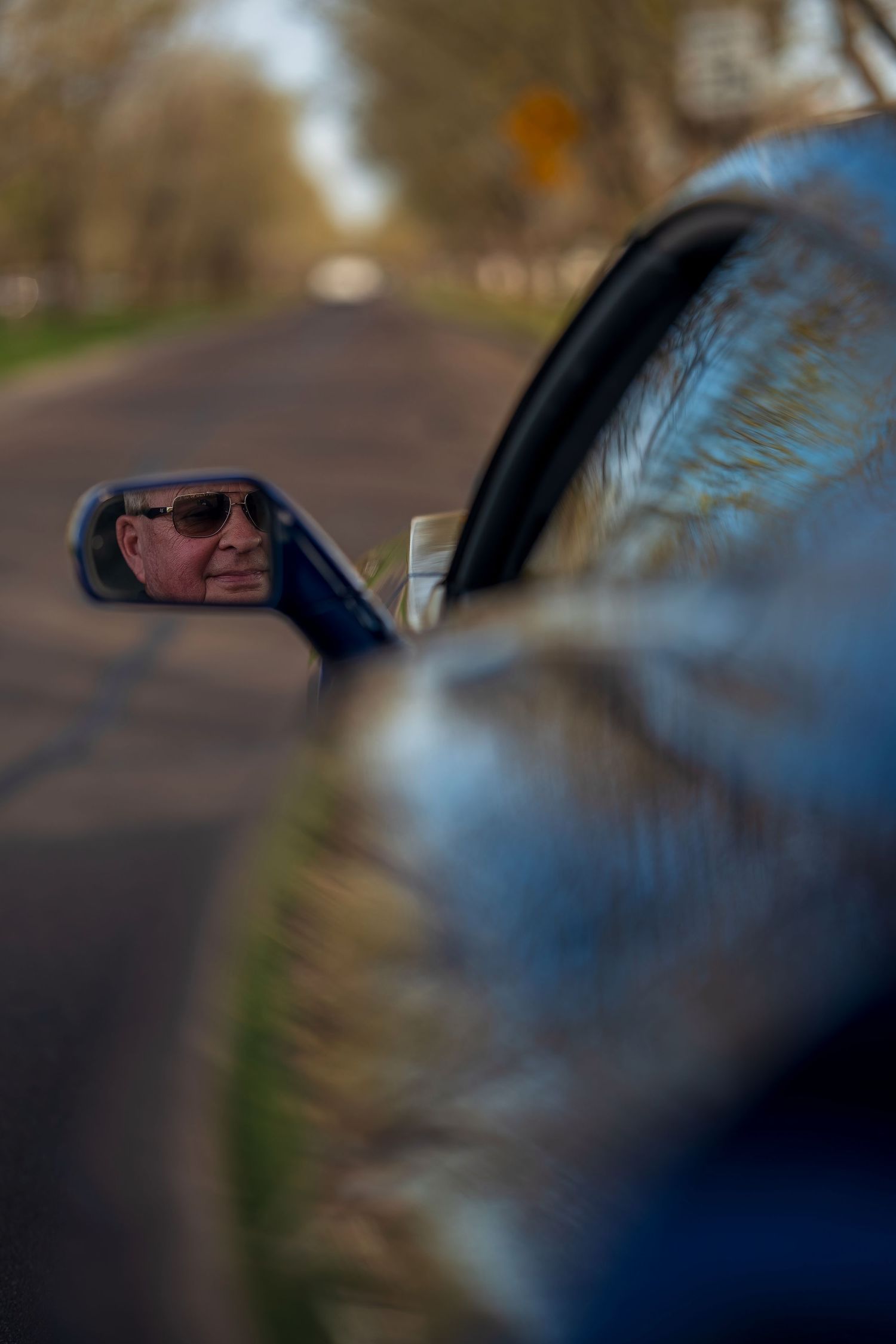 Reflection of an older man in sunglasses visible in the side mirror of a blue Corvette on a quiet road.