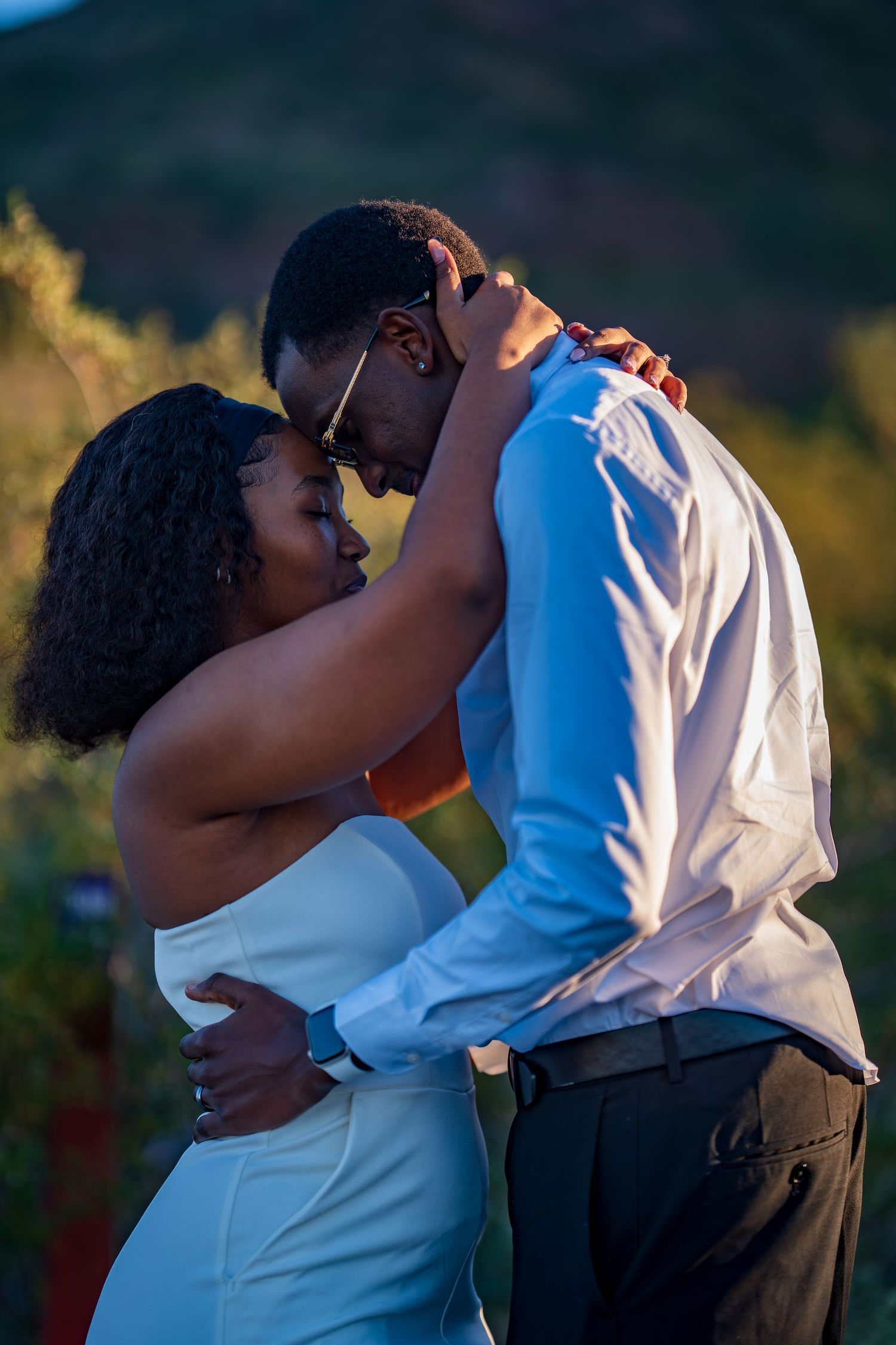 A couple shares a romantic embrace outdoors at sunset, with the woman in a white dress and man in a white shirt.