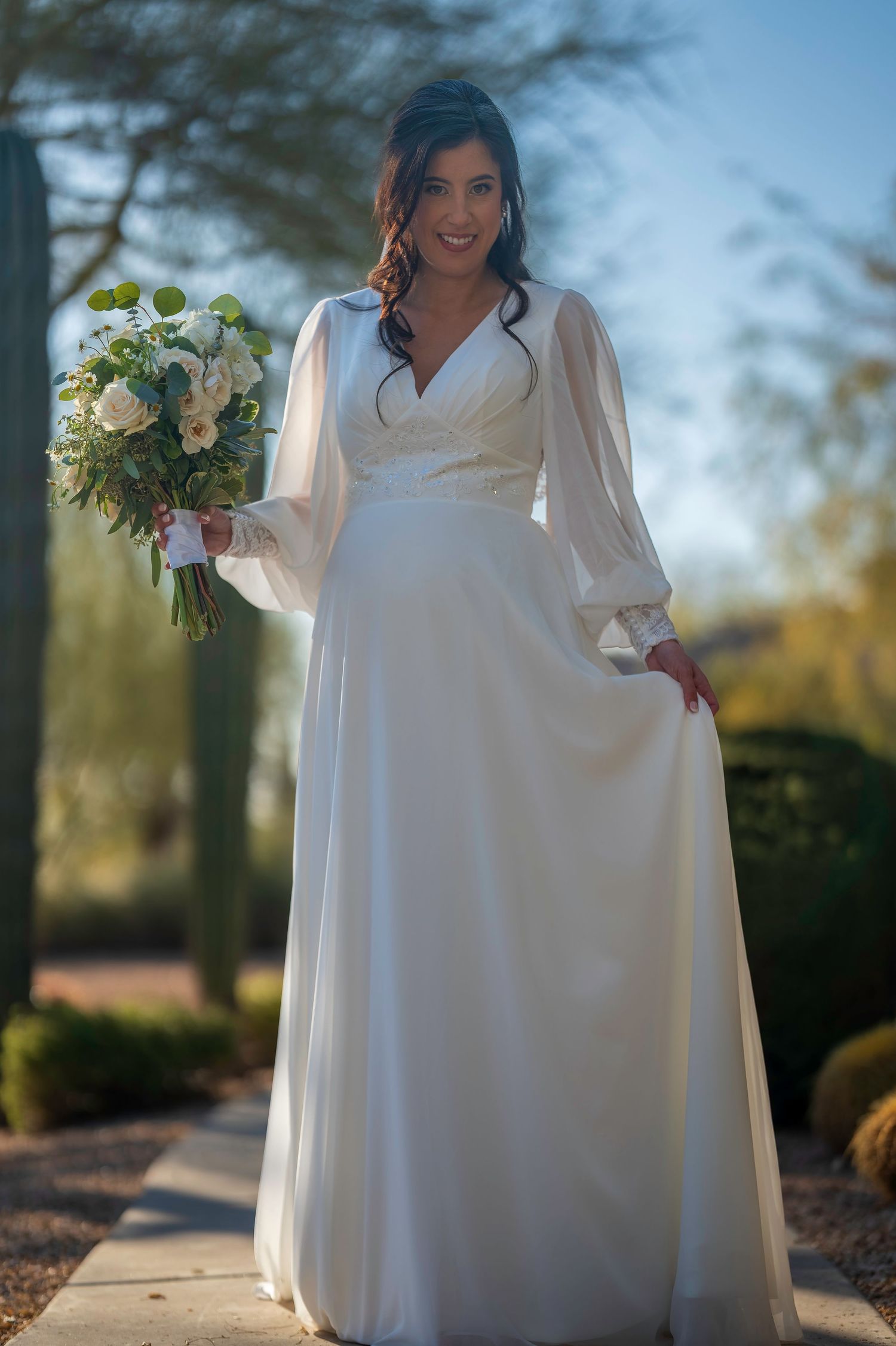 Bride in elegant white long-sleeve wedding gown holds bouquet of white and green flowers outdoors.